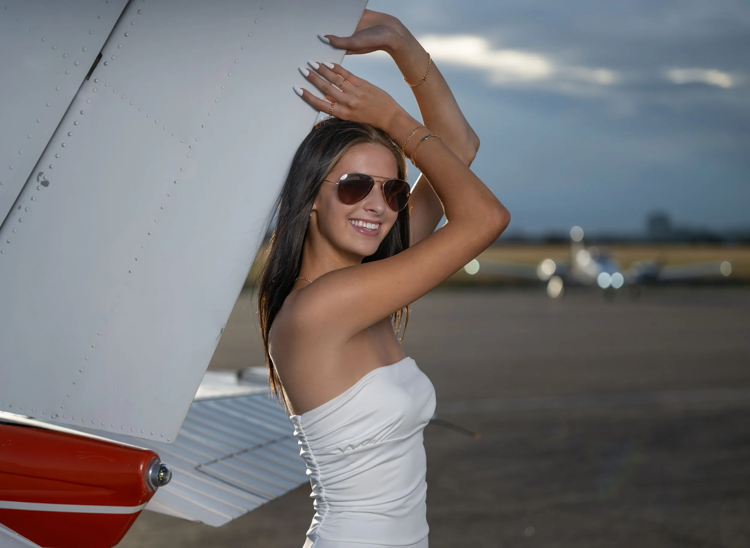 A woman in sunglasses and a white strapless dress standing next to an aircraft at an airport during sunset, smiling with one hand resting on the plane and the other raised above her head.