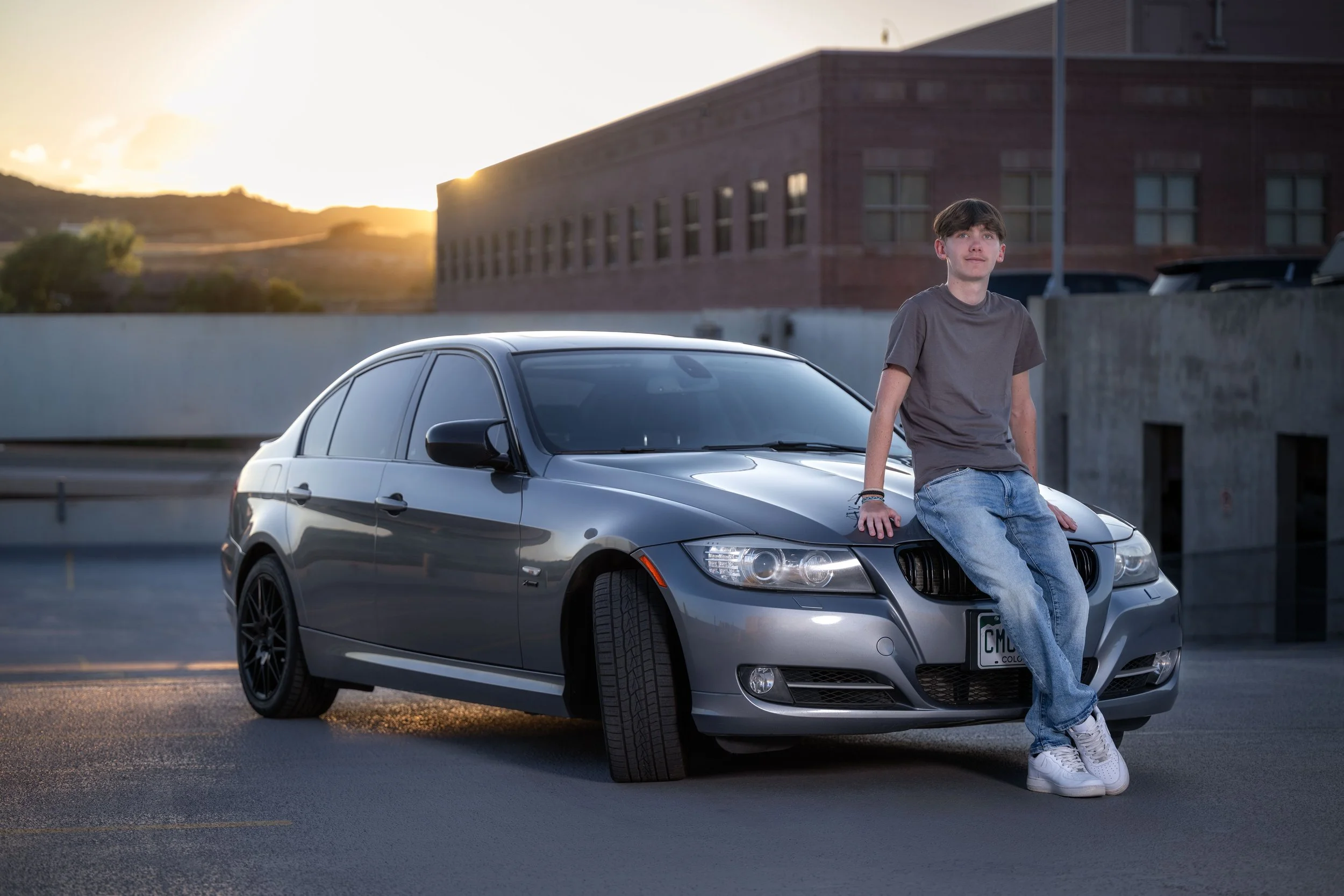 Young man in casual clothes sitting on the hood of a silver sedan car in an empty parking lot during sunset.