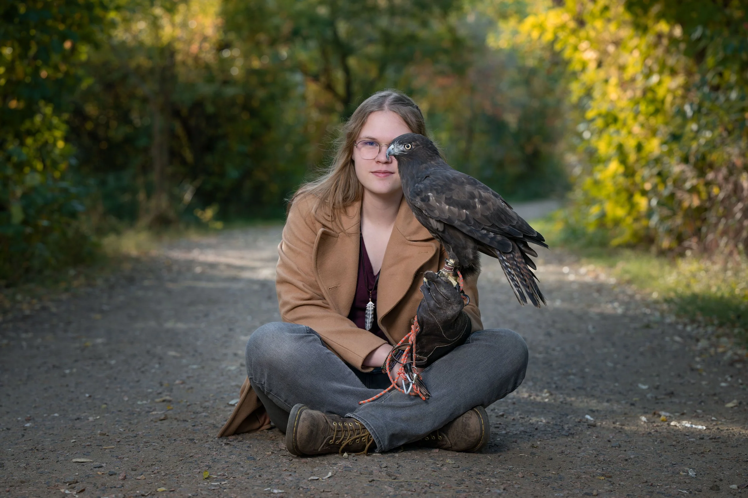 A woman sitting cross-legged on a dirt path in a forested area, holding a large bird of prey on her gloved hand. The woman has long blonde hair, glasses, and is wearing a tan coat and gray pants with brown boots. The bird, possibly a hawk or an eagle