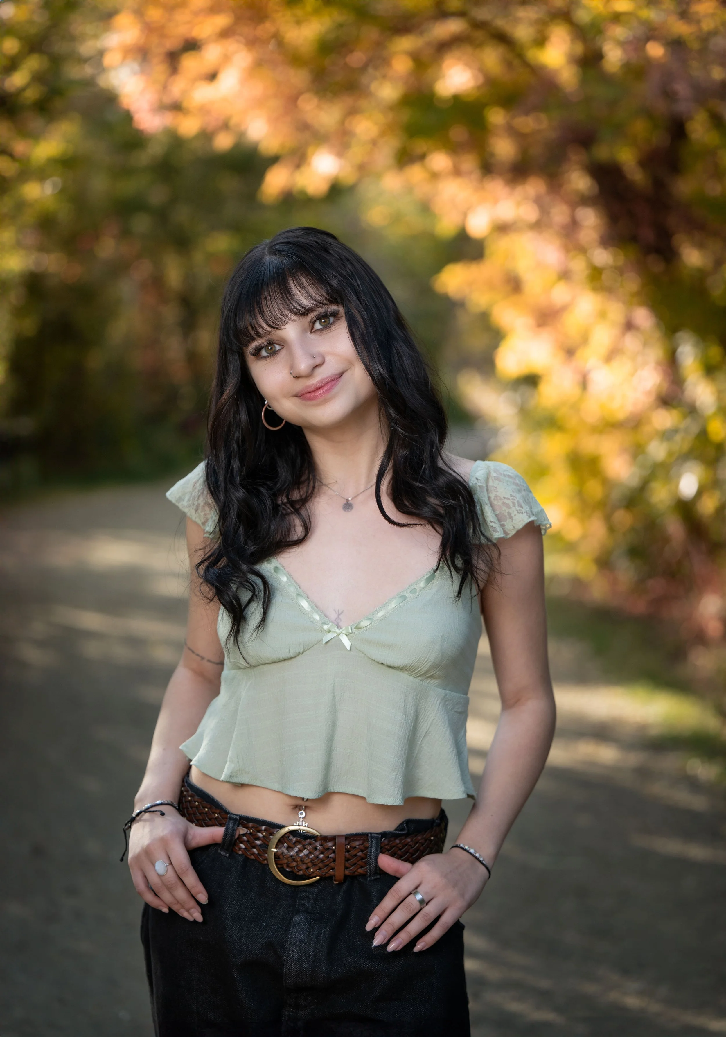 A young woman with dark hair and bangs standing outdoors on a dirt path, surrounded by colorful autumn trees, smiling at the camera.