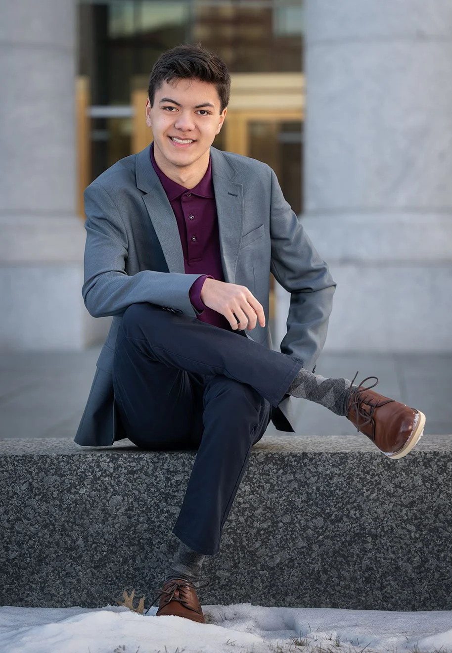 A young man in a gray blazer and maroon shirt sitting on a stone ledge outdoors, with a modern building in the background and snow on the ground.