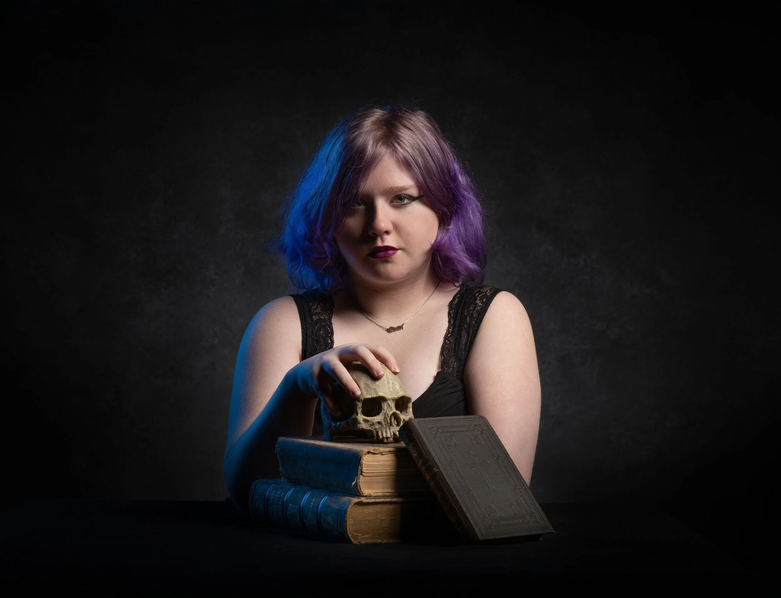 A woman with purple and blue hair sits at a table with old books, holding a small skull, against a dark background.