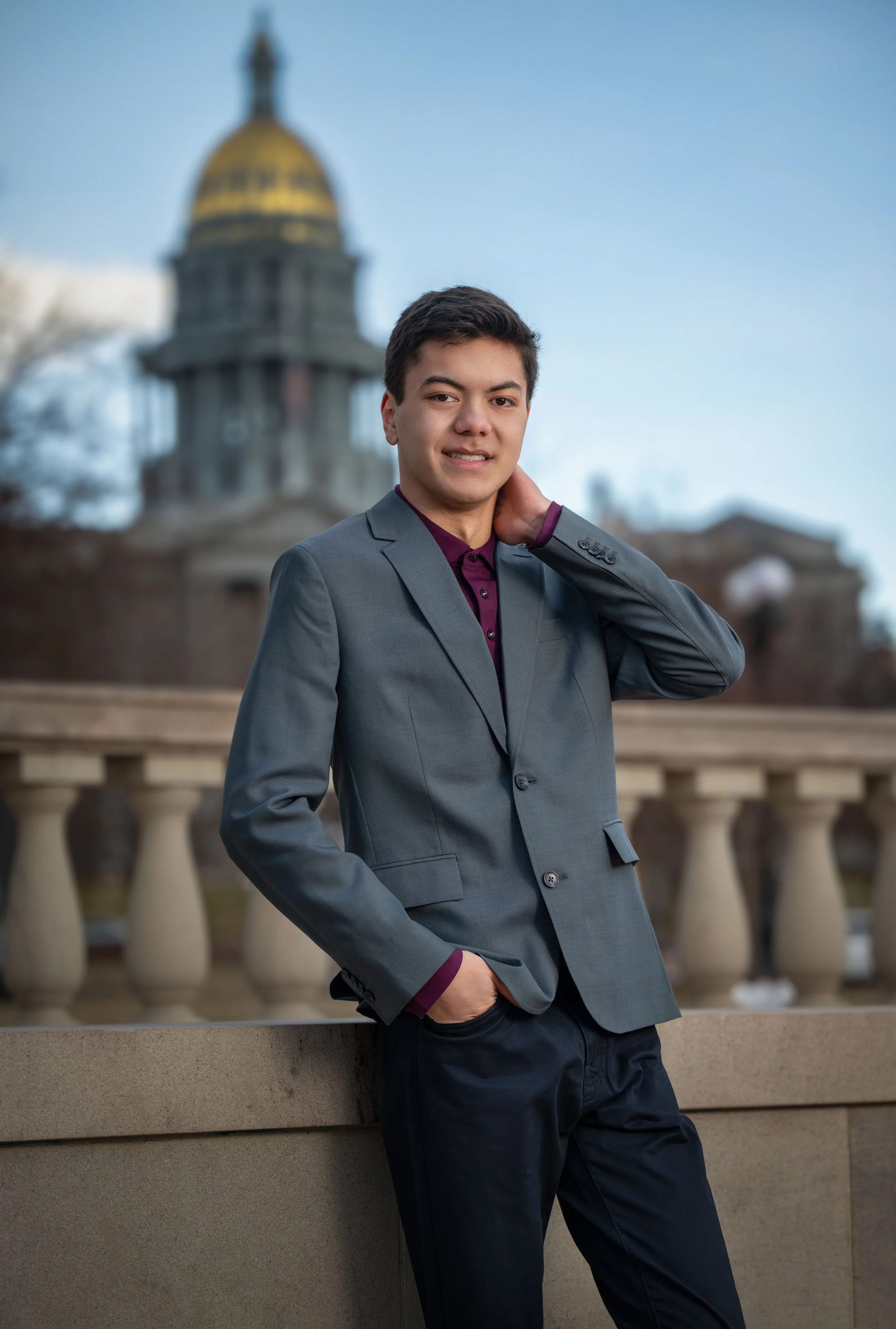 A young man in a gray blazer and dark pants standing outdoors in front of a stone railing with a domed building in the background.