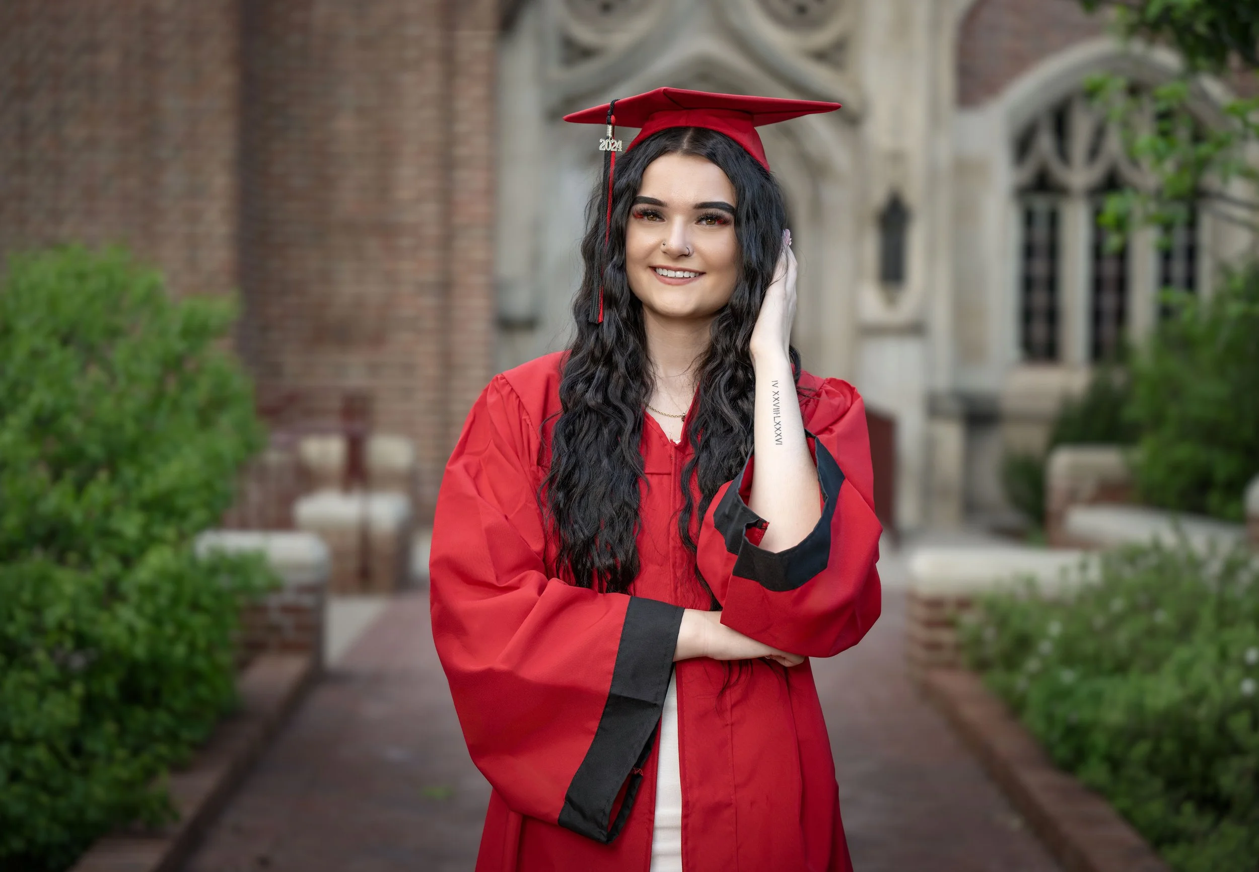 A young woman in a red graduation gown and cap standing outdoors in front of a historic brick building with greenery, smiling at the camera.