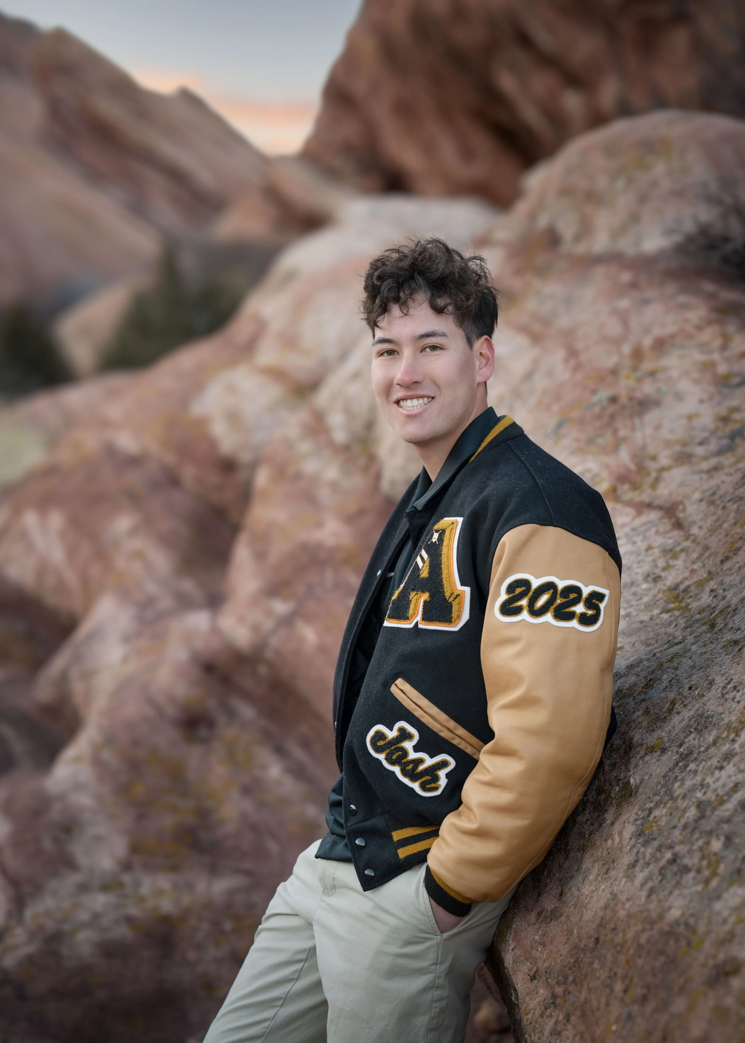 A young man standing outdoors against large rocks, wearing a black and beige varsity jacket with the year 2025 on the sleeve, smiling and looking at the camera.