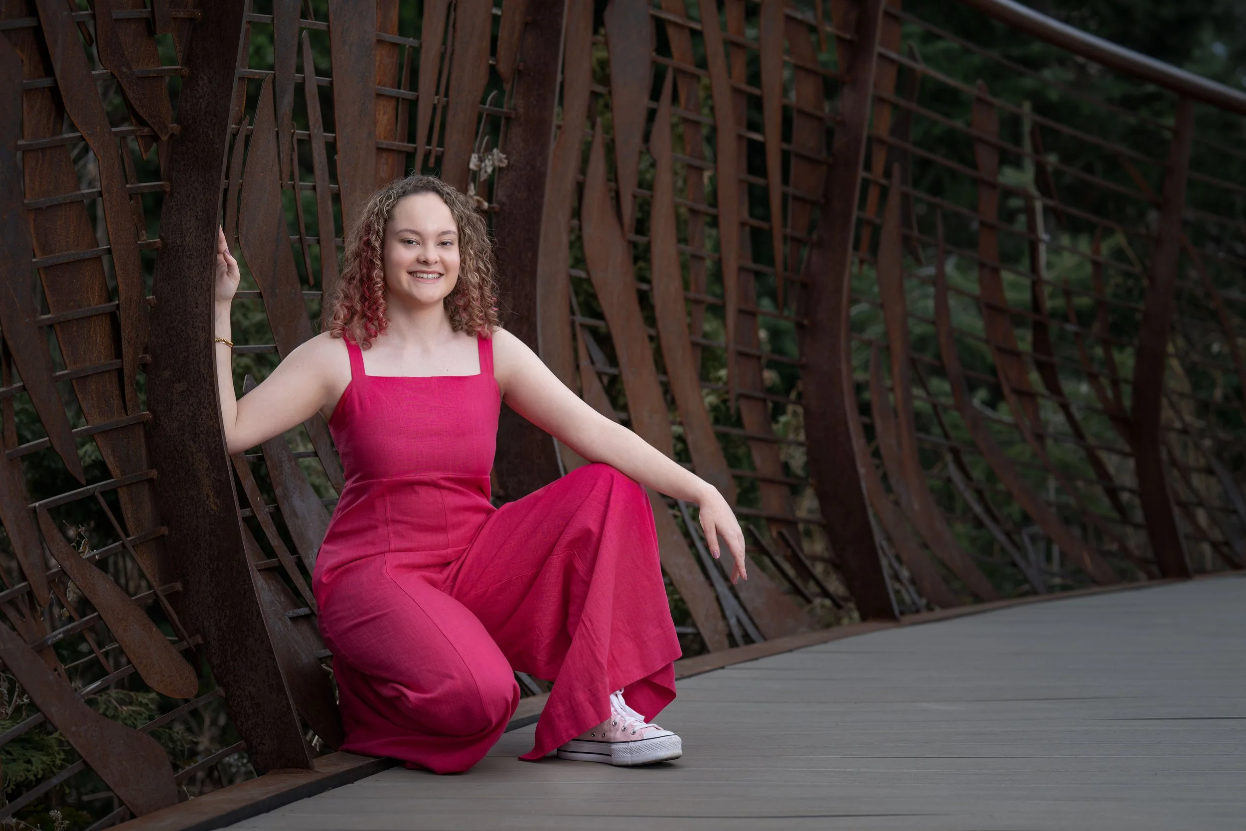 A young woman with curly hair and pink highlights sitting on a curved wooden bridge. She is wearing a bright pink jumpsuit and white sneakers, smiling at the camera. The background shows a metal structure and green foliage.