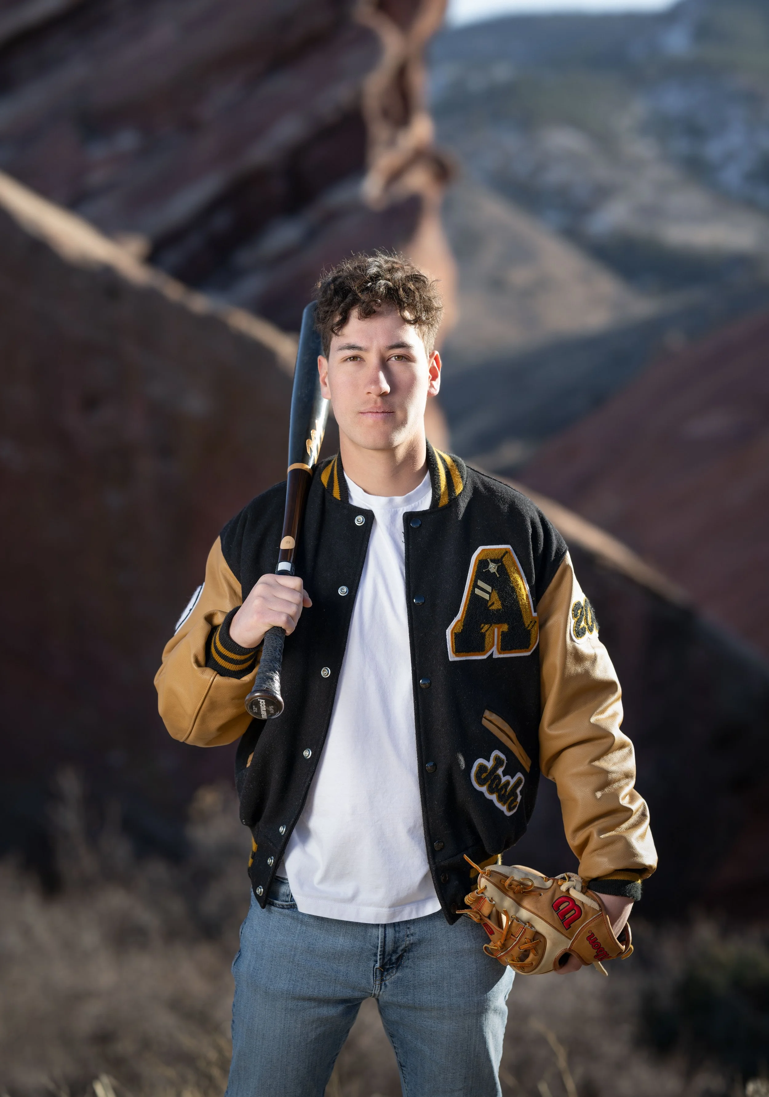 A young man holding a baseball bat over his shoulder, wearing a black and tan varsity jacket with patches, a white t-shirt, and jeans, standing outdoors with a mountainous landscape in the background.