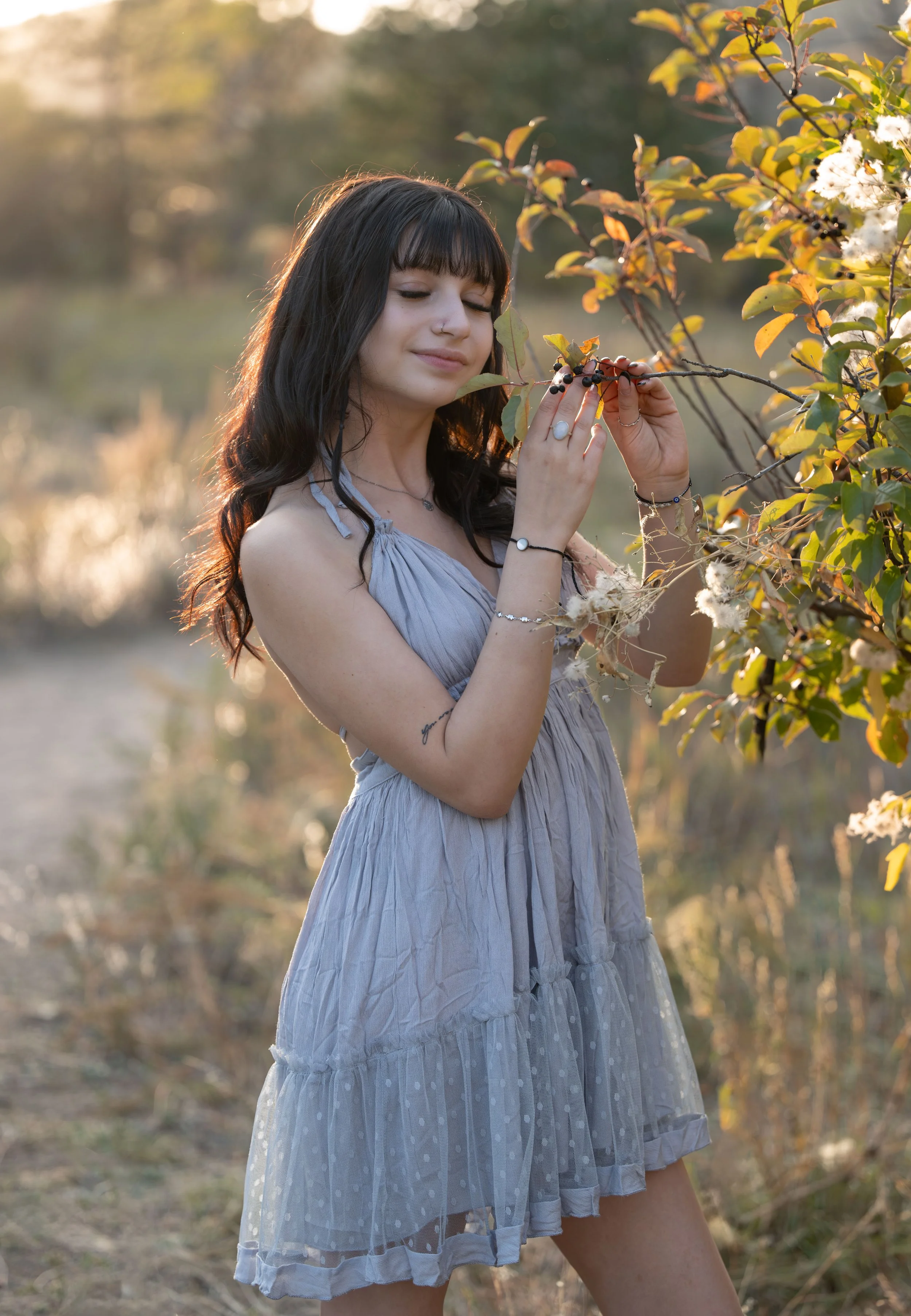 A young woman with dark hair and bangs standing outdoors near water, gently holding a small branch with black berries and white flowers, illuminated by golden sunlight.