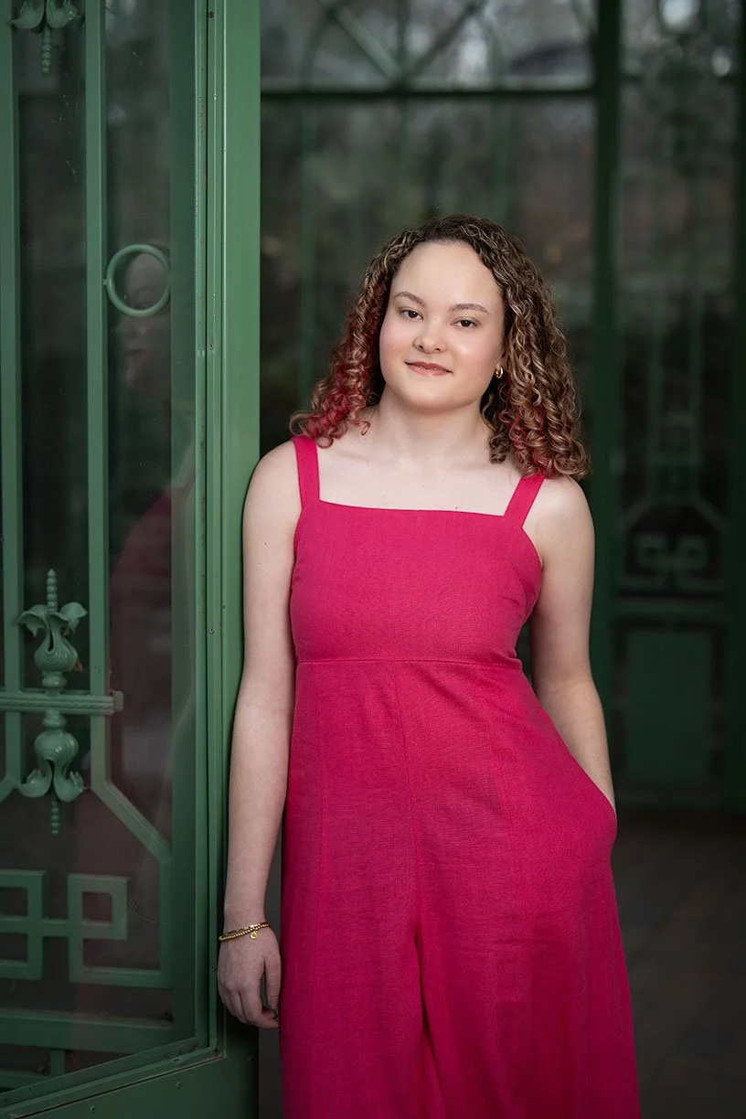 A young woman with curly hair wearing a pink sleeveless dress standing next to a glass door frame.