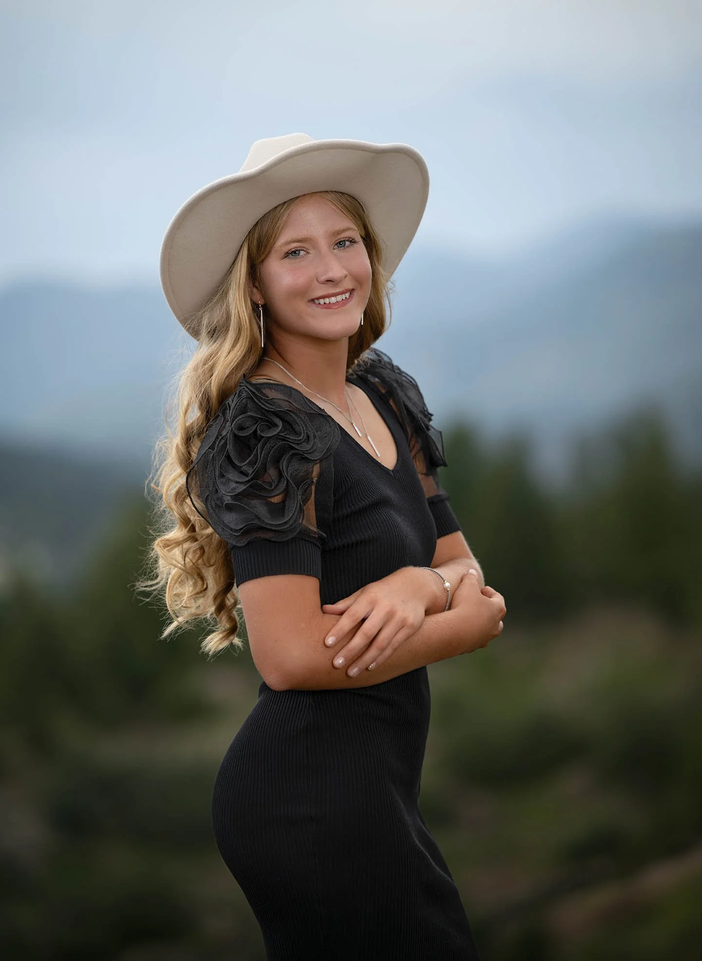 Young woman with long curly blonde hair wearing a wide-brimmed white hat, black dress with ruffled sleeves, standing outdoors with a blurred natural background.