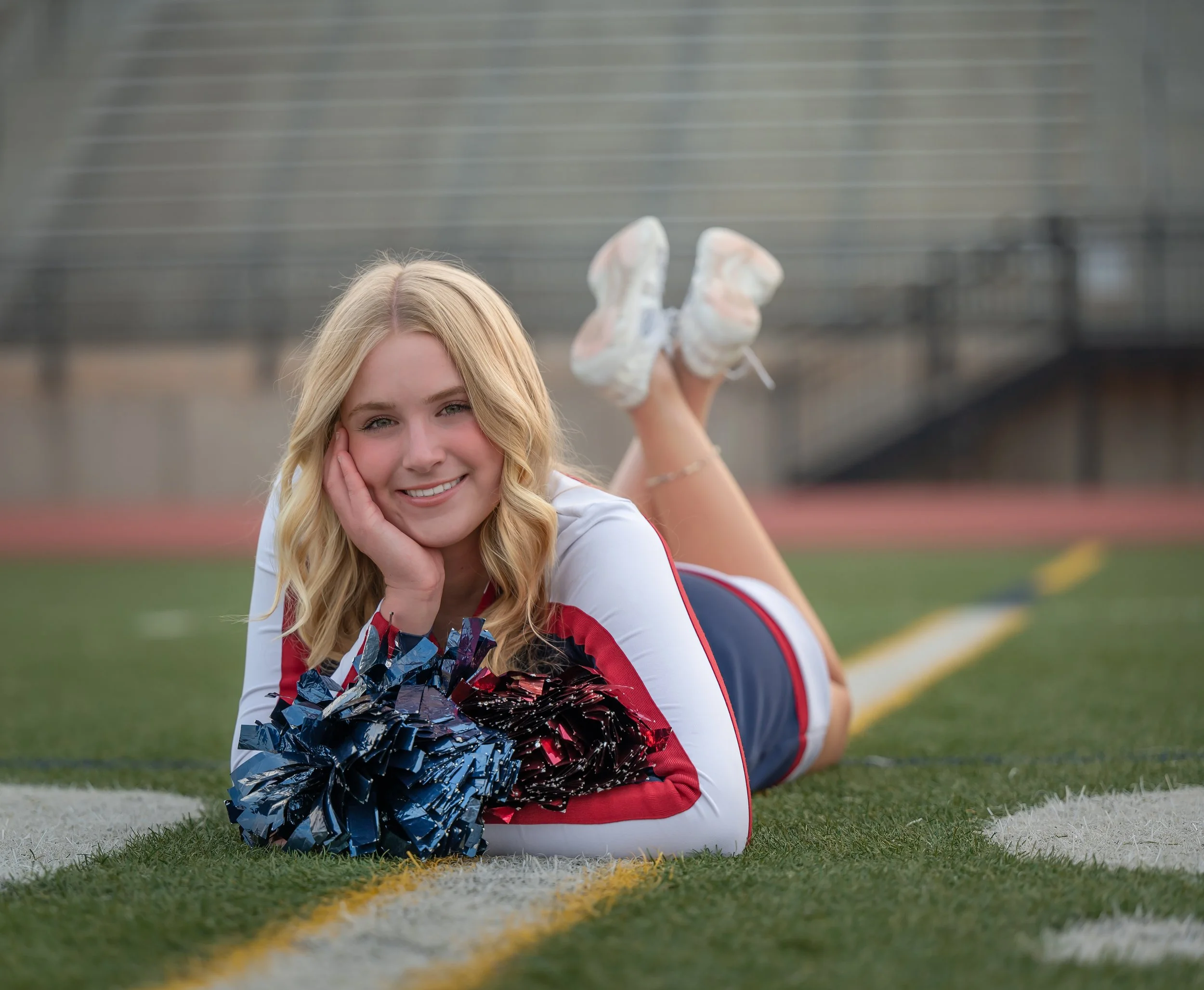 A young woman with blonde hair lying on her stomach on a football field, holding red and blue pom-poms, with her feet raised in the air.