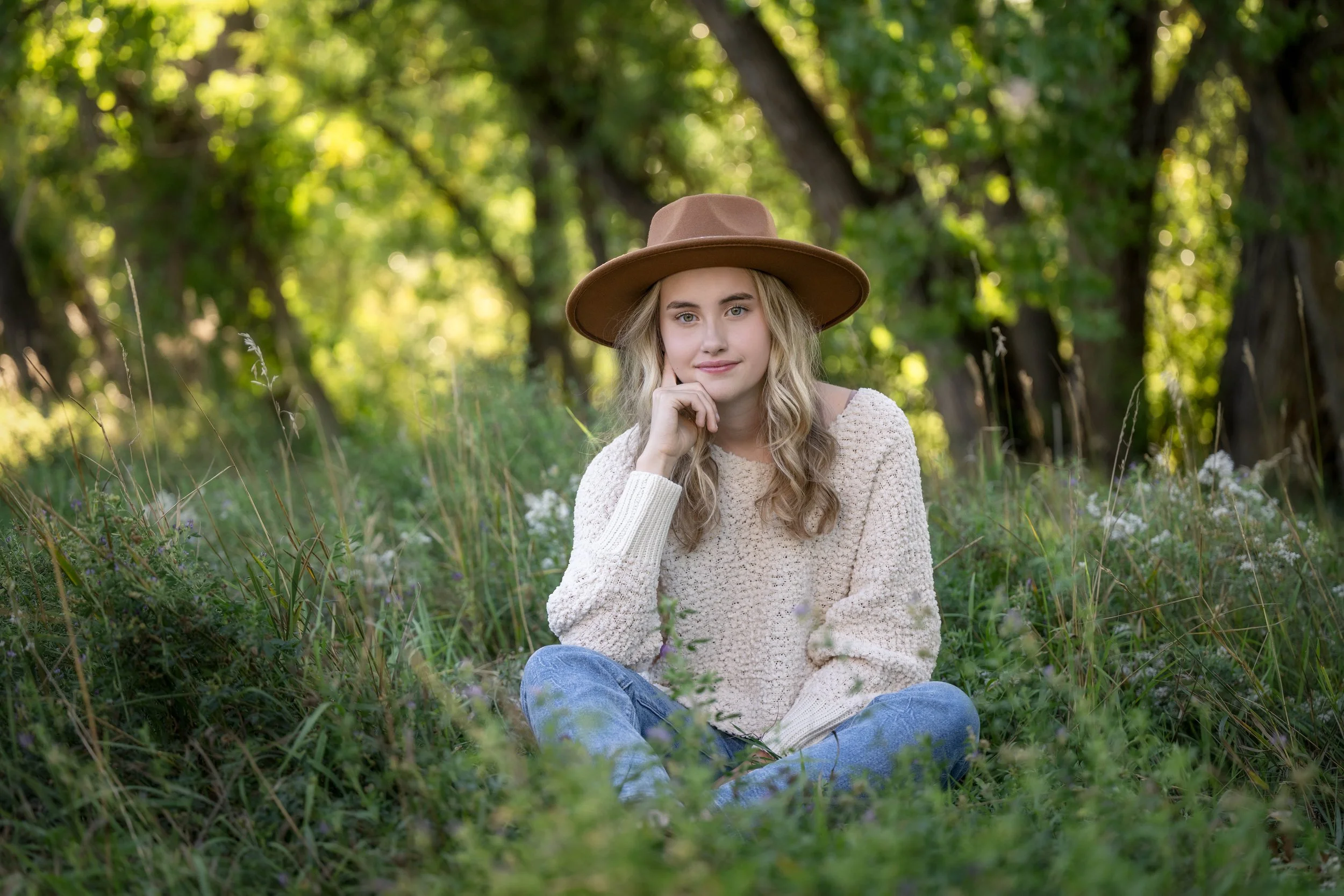 A young woman with blonde hair, wearing a beige sweater and a brown wide-brimmed hat, sitting cross-legged on the grass in a wooded area, resting her chin on her hand, surrounded by greenery and trees.