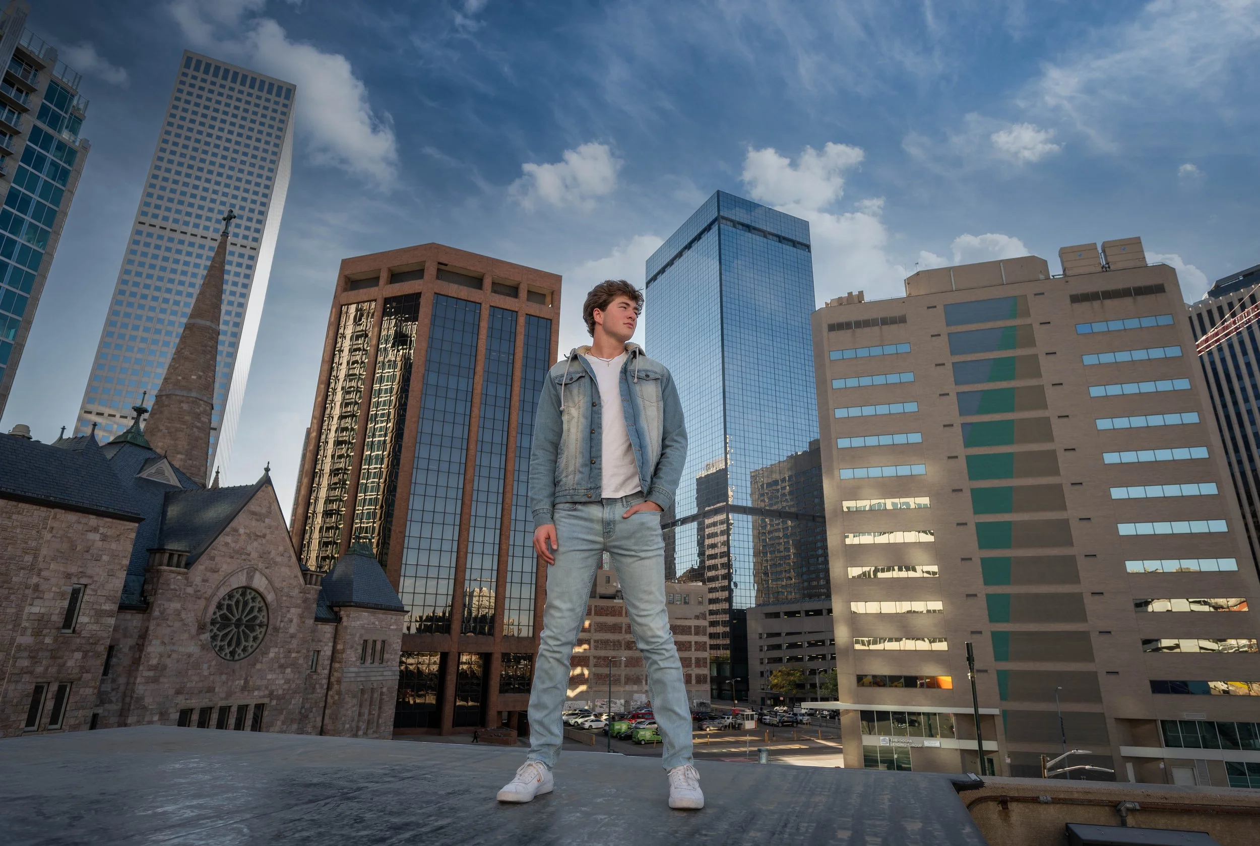 Teenage boy in light denim jacket and jeans standing on rooftop with city skyscrapers in the background.