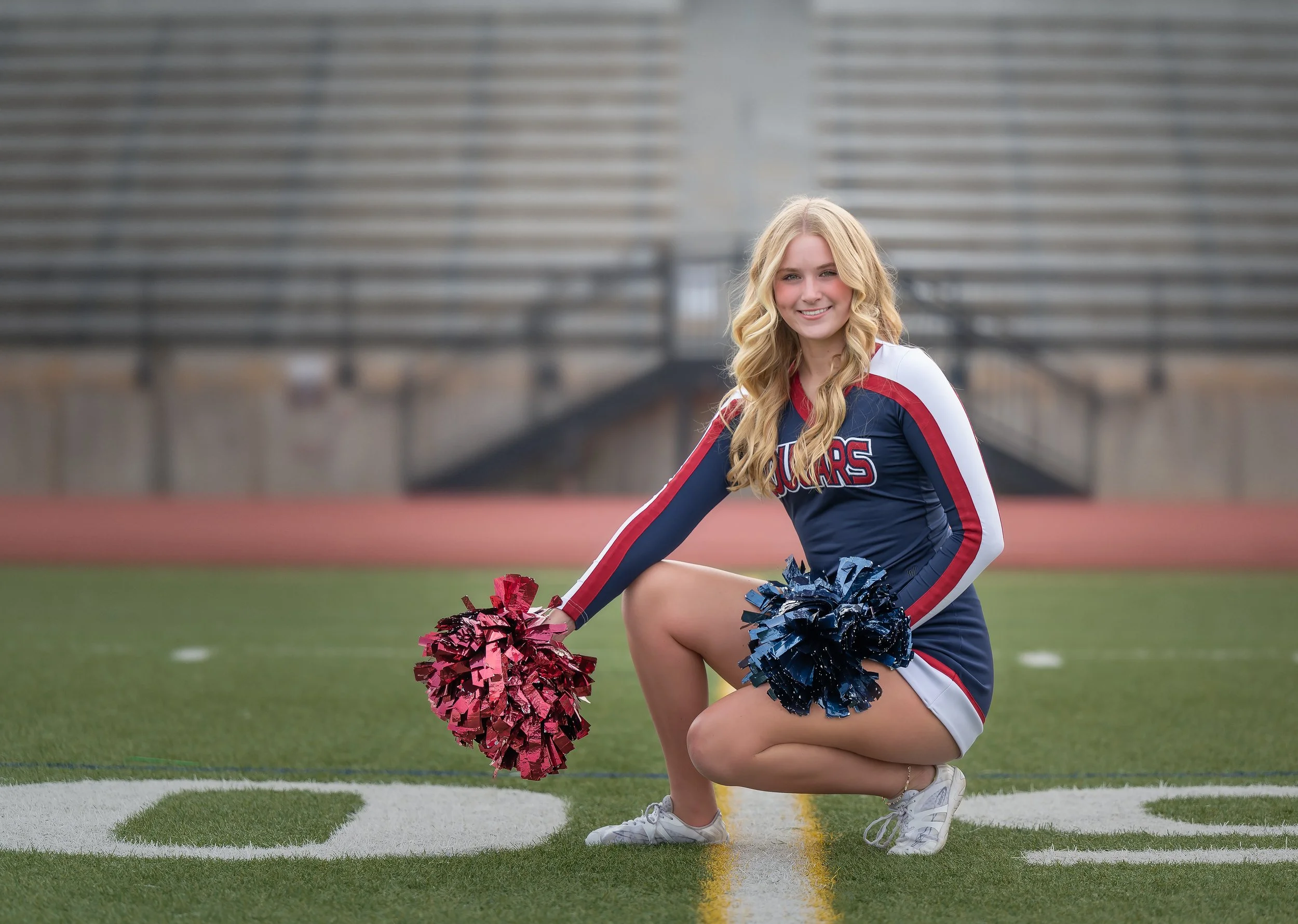 A young female cheerleader in a navy, red, and white uniform kneels on a football field, holding pink and navy pom-poms, with bleachers in the background.