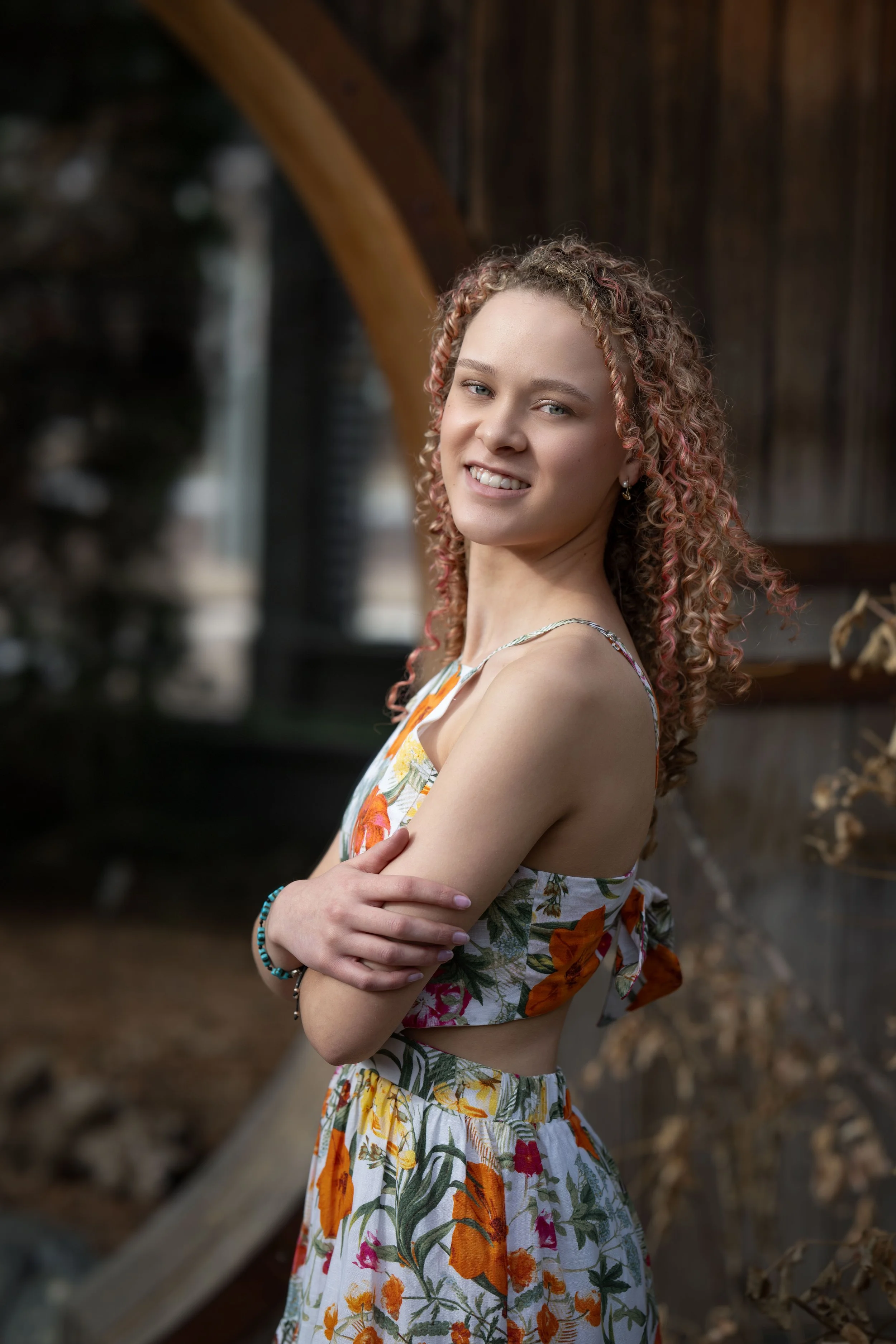 A young woman with curly hair in a floral dress, smiling and crossing her arms, standing outdoors.