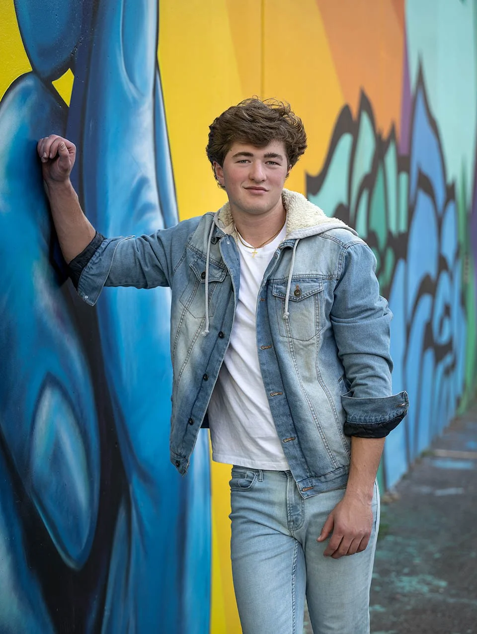A young man with curly brown hair wearing a denim jacket, white t-shirt, and jeans, leaning against a colorful graffiti wall with his left arm raised and hand clenched in a fist.