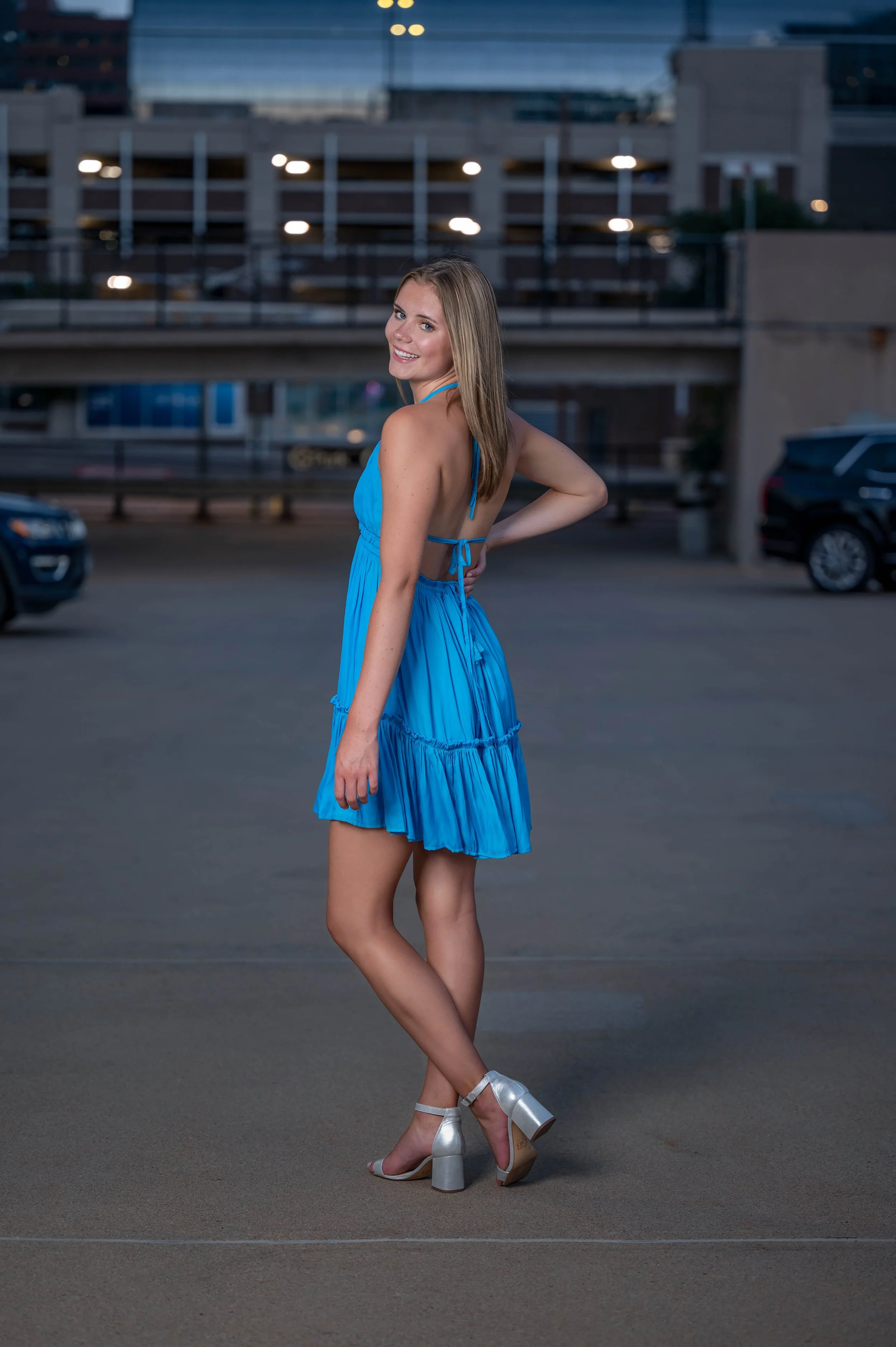 A young woman in a bright blue dress with open back and silver high heel sandals standing on a parking lot during dusk, smiling and posing.