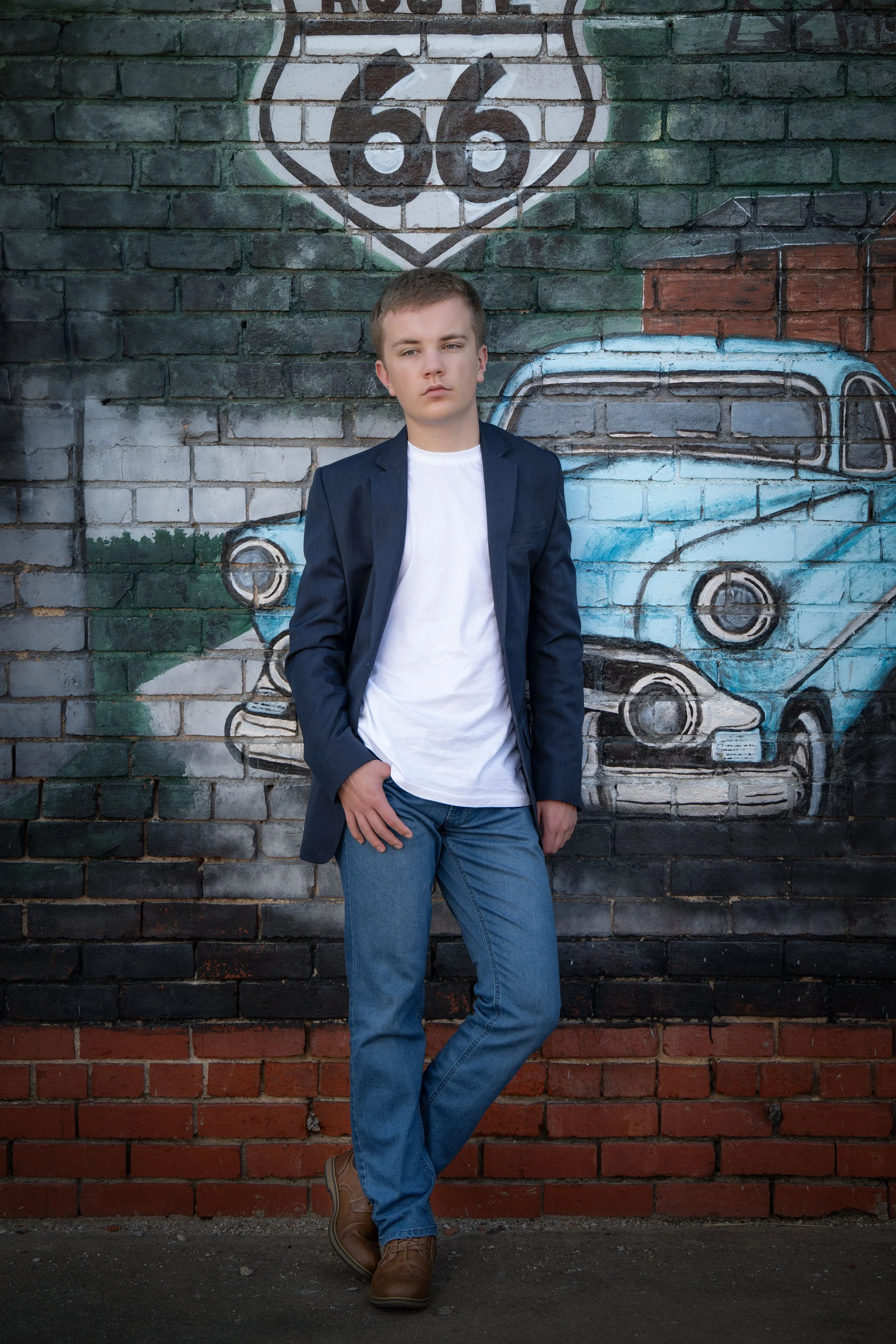 A young man in a white T-shirt, blue blazer, and jeans standing against a brick wall with urban graffiti, including a vintage car and a Route 66 sign.