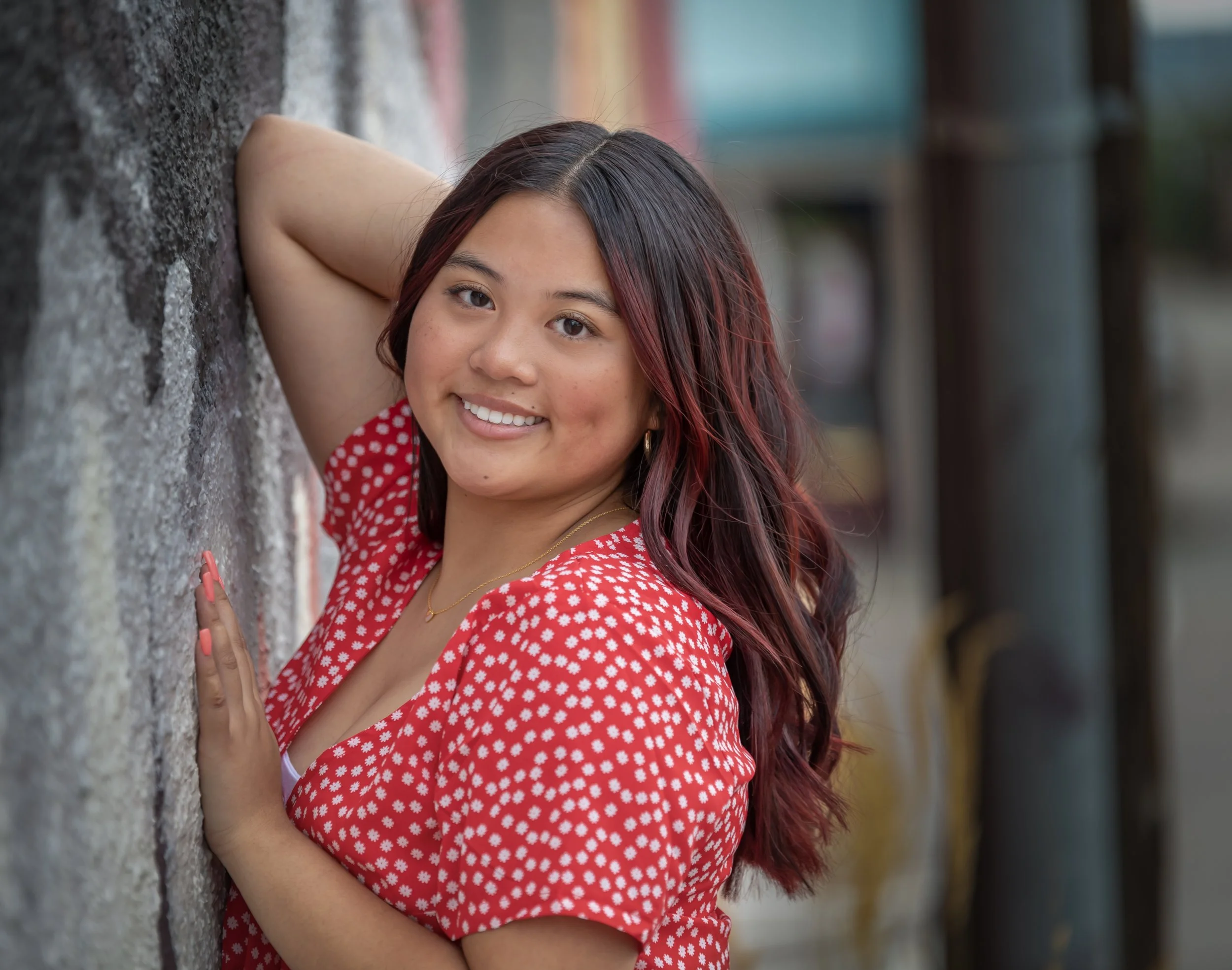 A young woman with long, wavy dark hair with reddish highlights, smiling, wearing a red and white polka dot dress, leaning against a textured stone wall