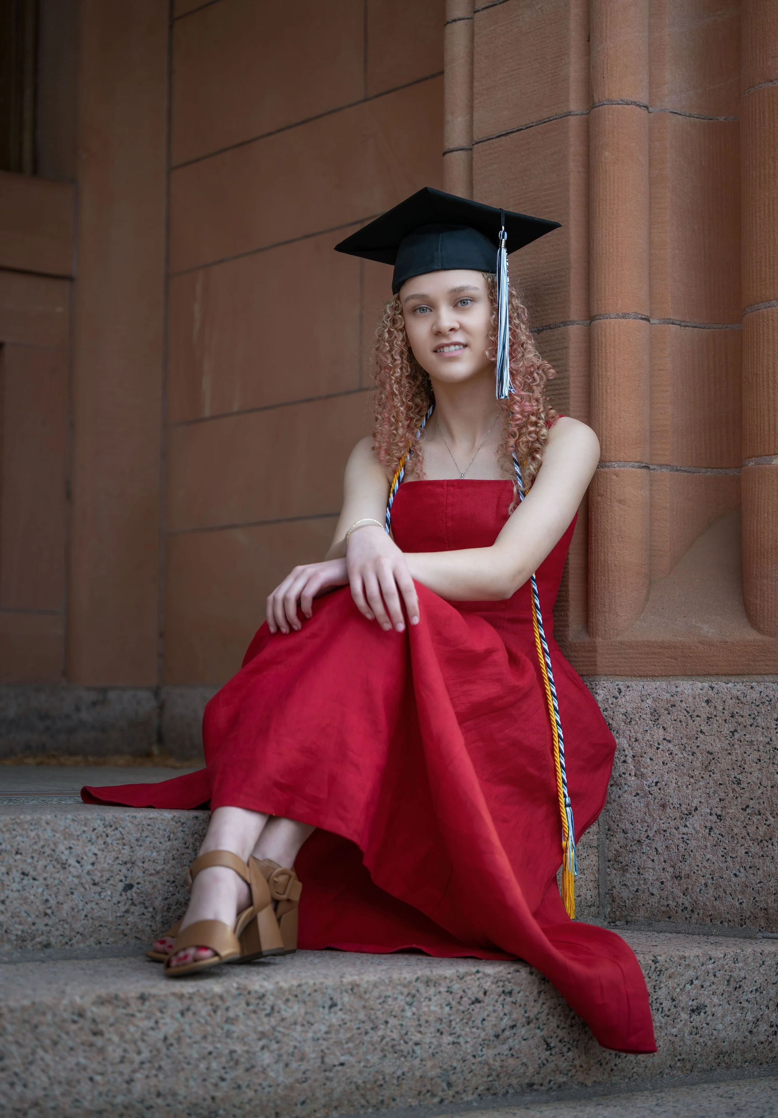 Young woman in a red dress sitting on steps outside a building, wearing a graduation cap and cords.