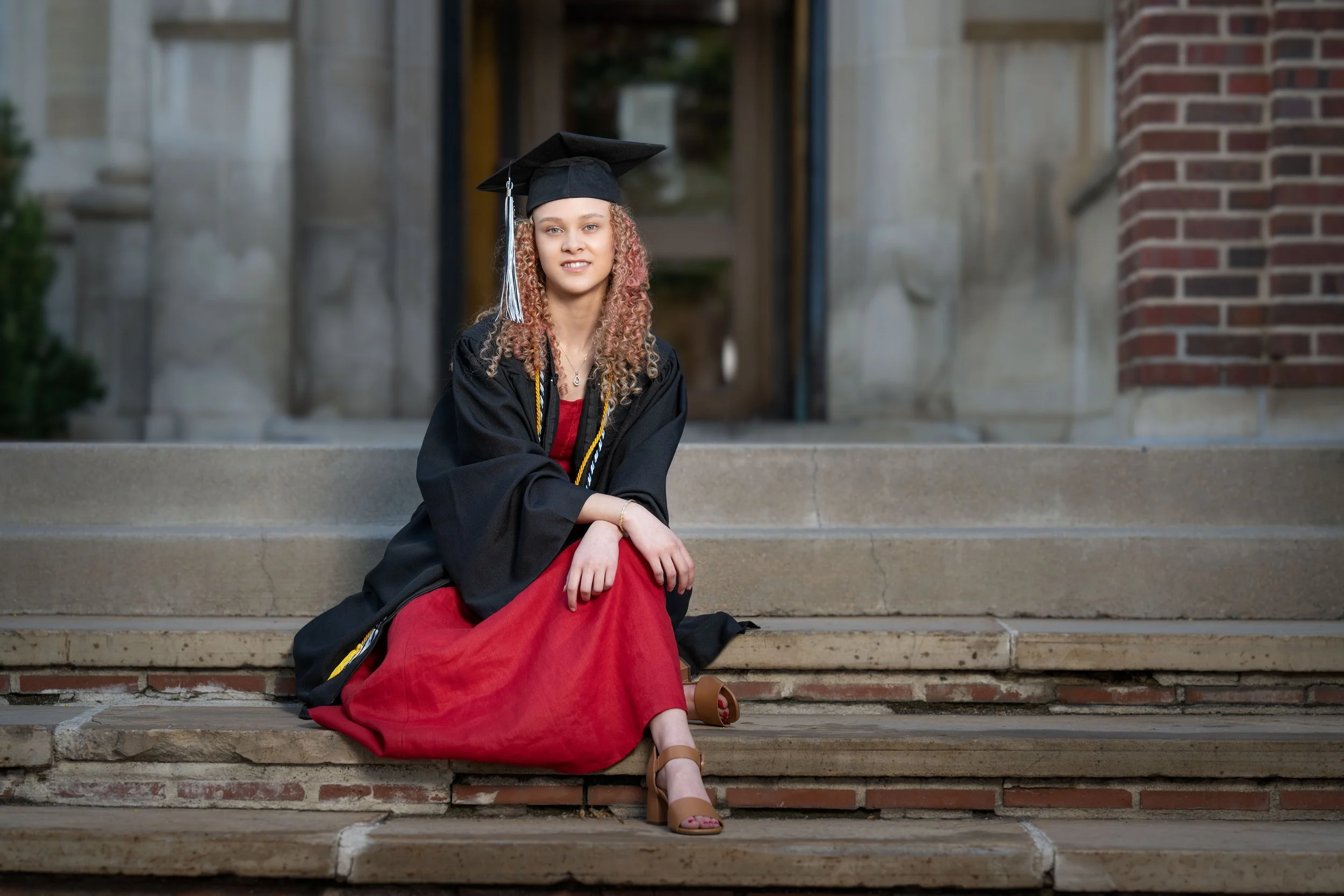 A young woman in a black graduation cap and gown sitting on stone steps outside, wearing a red dress and brown high-heeled shoes, celebrating graduation.
