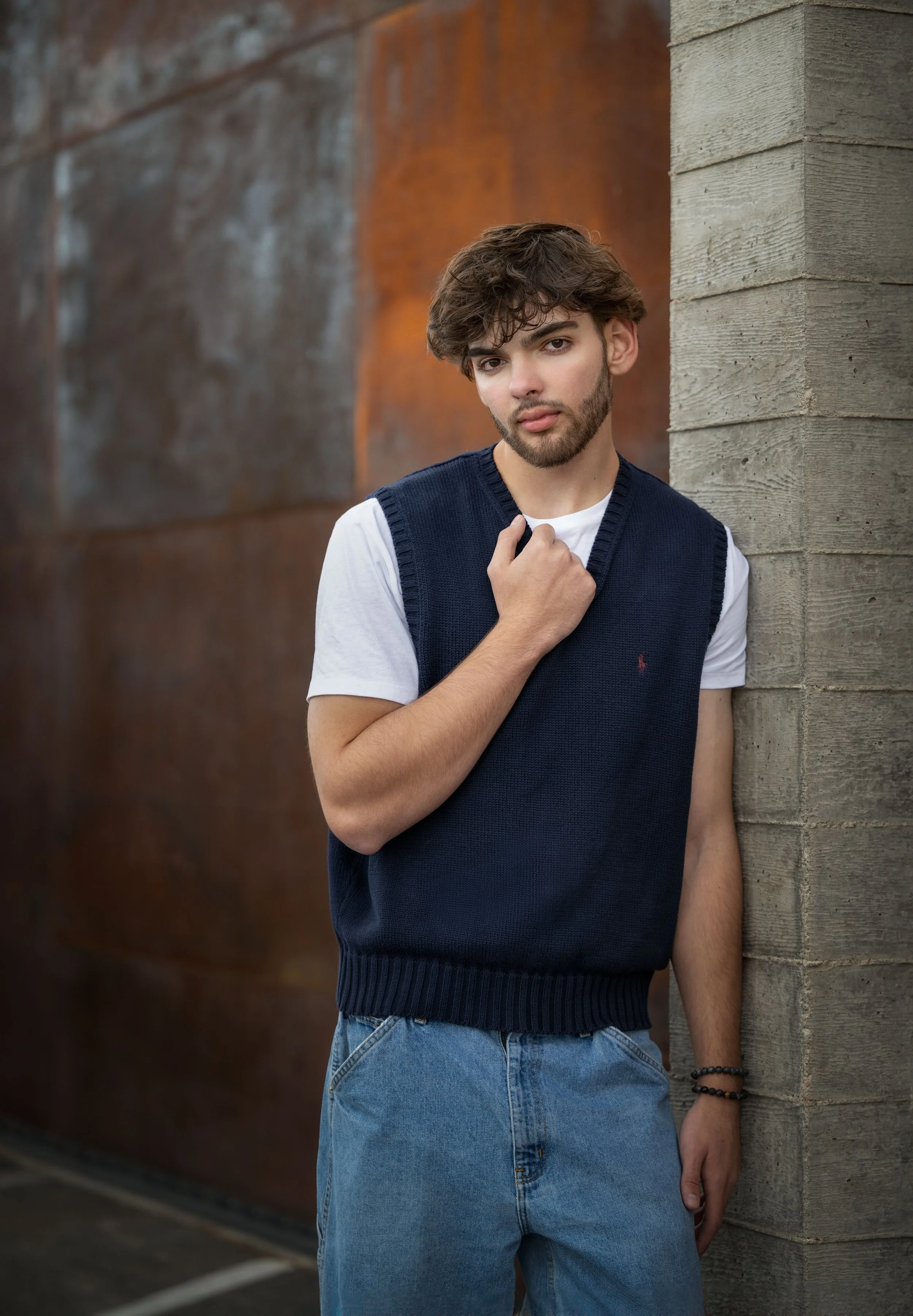 A young man with brown hair and a beard is standing by a concrete wall and a rusted metal wall. He is wearing a white t-shirt with a navy blue sleeveless sweater vest, and light blue jeans. He has a bracelet on his left wrist and is touching his ches