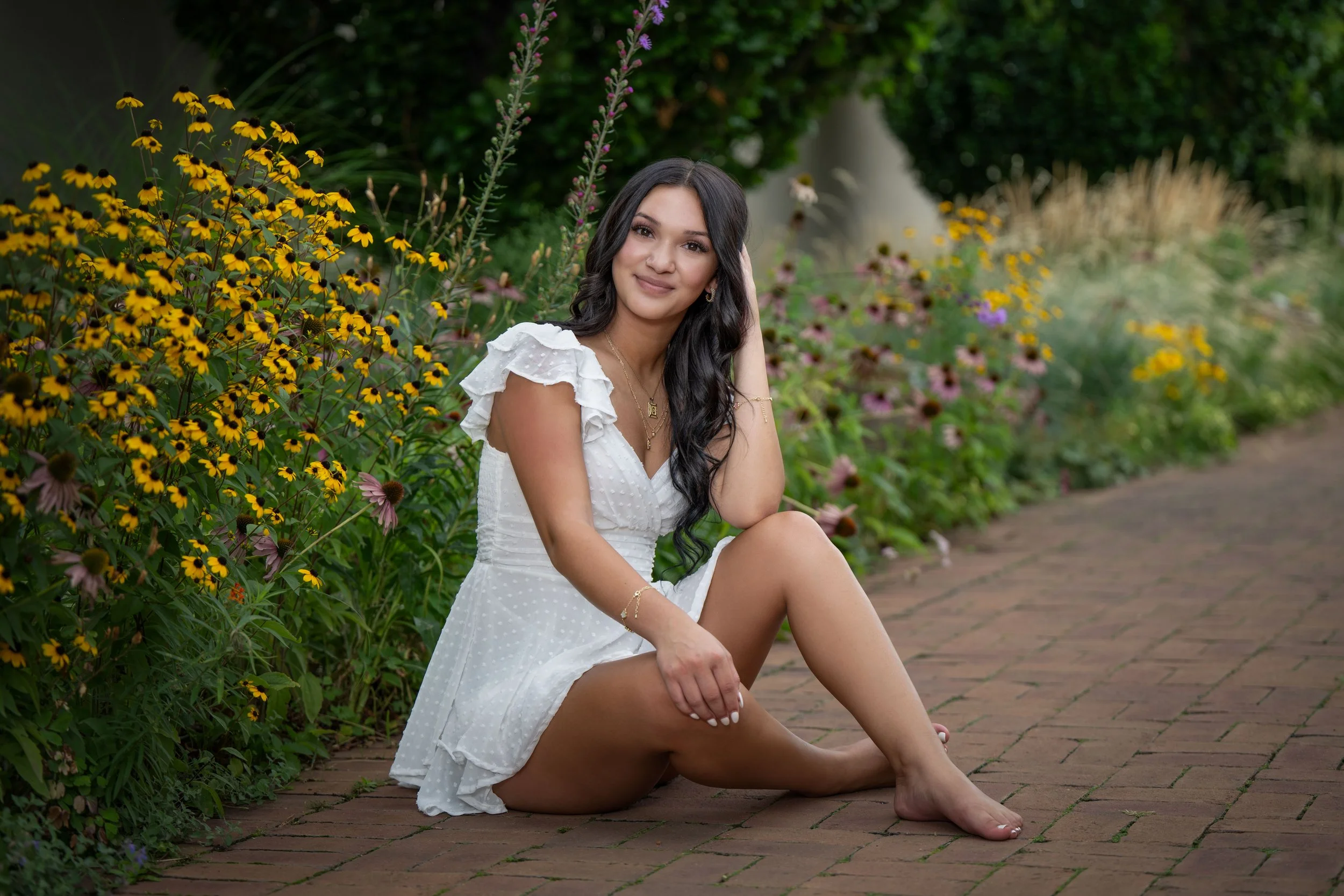 Natural senior portraits in a white dress sitting barefoot on a brick path amidst colorful flowers in a garden.