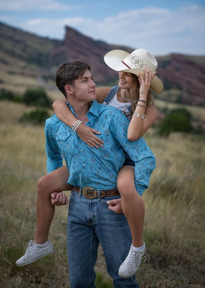 A young man carries a young woman on his shoulders in a field with mountains in the background. The woman is smiling and adjusting her wide-brimmed cowboy hat, with her arm around the man's neck. The man is looking at her with a neutral expression, w