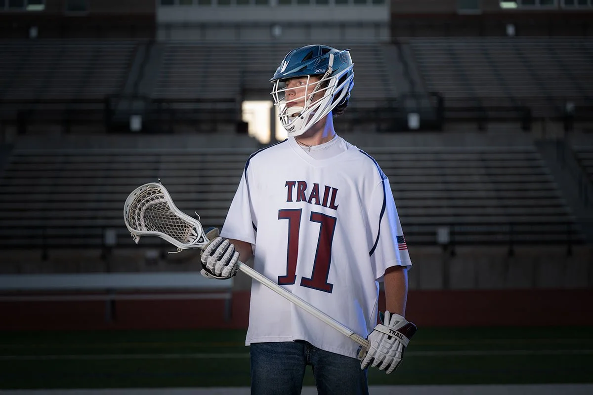 A young male lacrosse player wearing a helmet, white jersey with 'TRAIL 11' on the front, holding a lacrosse stick, standing on an empty stadium field.