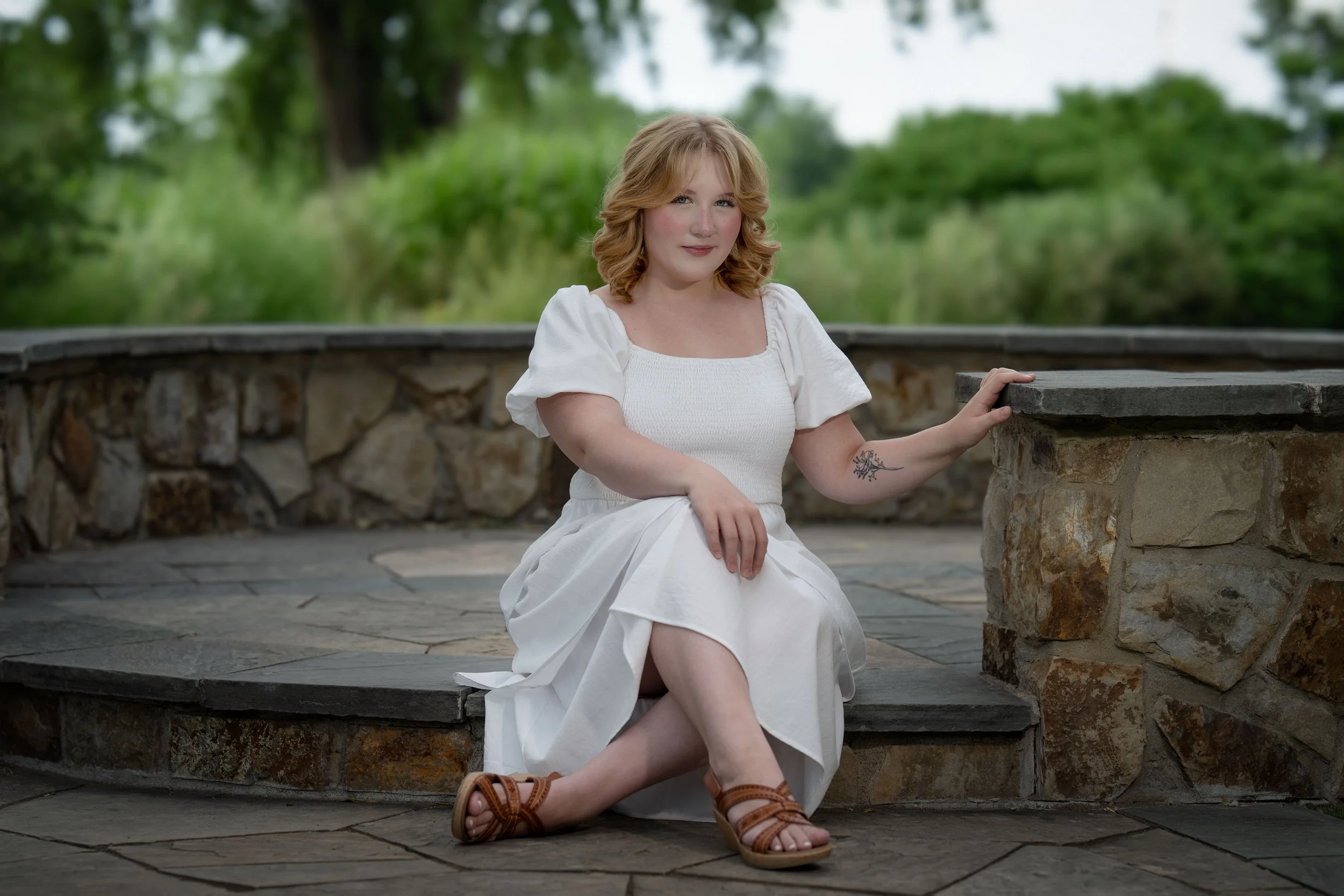 A young woman with shoulder-length curly blonde hair and light skin, wearing a white dress and brown sandals, sitting on stone steps outdoors with greenery in the background.