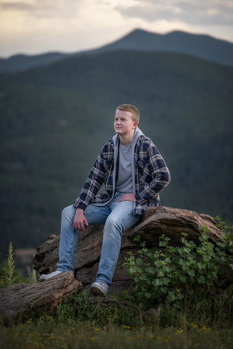 Young boy sitting on a large rock outdoors, dressed in a plaid jacket, gray t-shirt, and jeans, with mountains in the background during twilight.