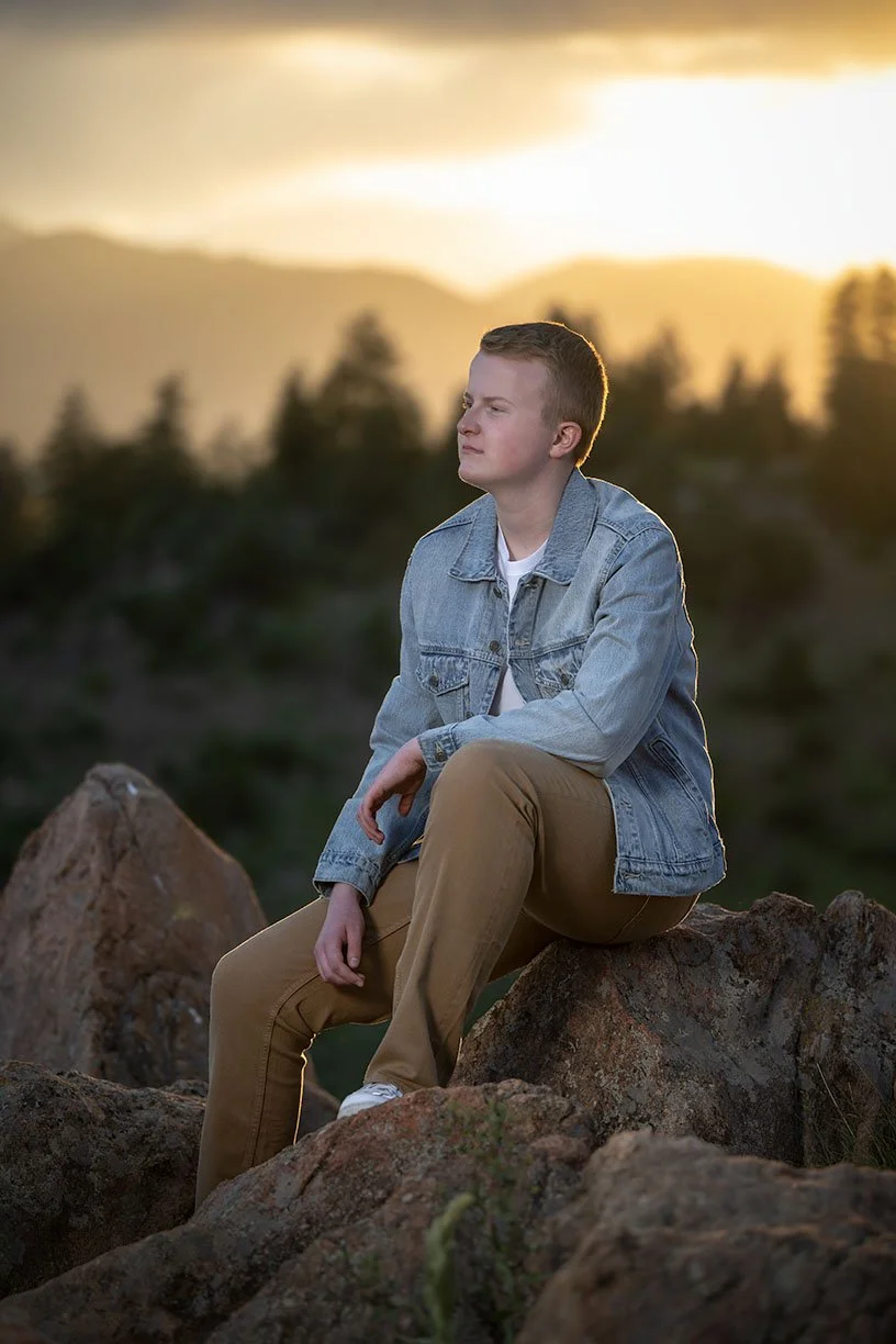 A young man sitting on rocks outdoors during sunset, wearing a denim jacket and khaki pants, looking thoughtfully into the distance.