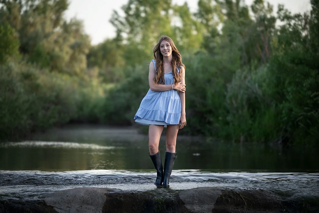 Young woman with long hair smiling while standing in a shallow river, wearing a blue dress and black rain boots, surrounded by green trees.