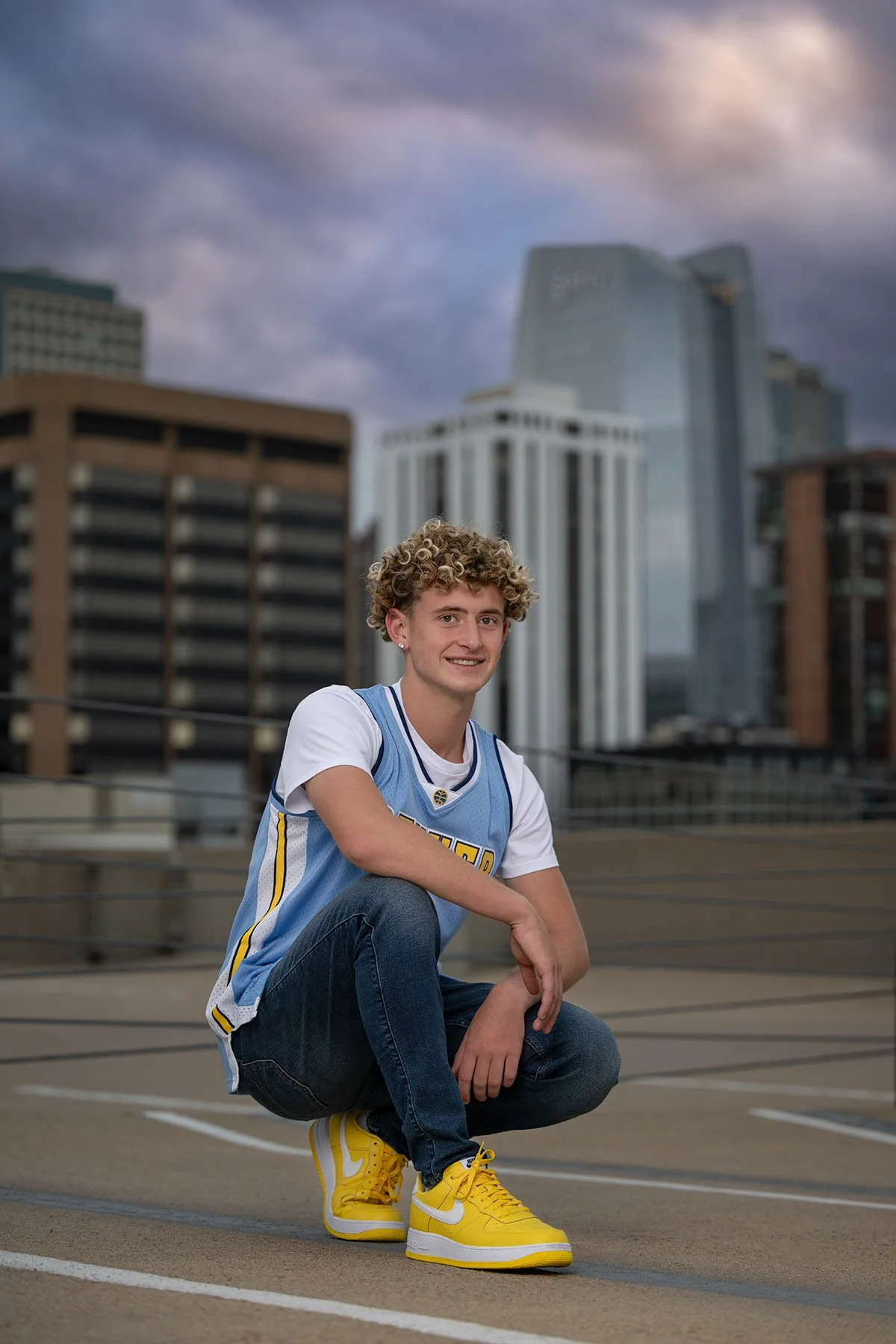 Young man with curly hair wearing a light blue and white sports jersey, jeans, and yellow sneakers, crouching on a rooftop parking lot with city buildings and a cloudy sky in the background.
