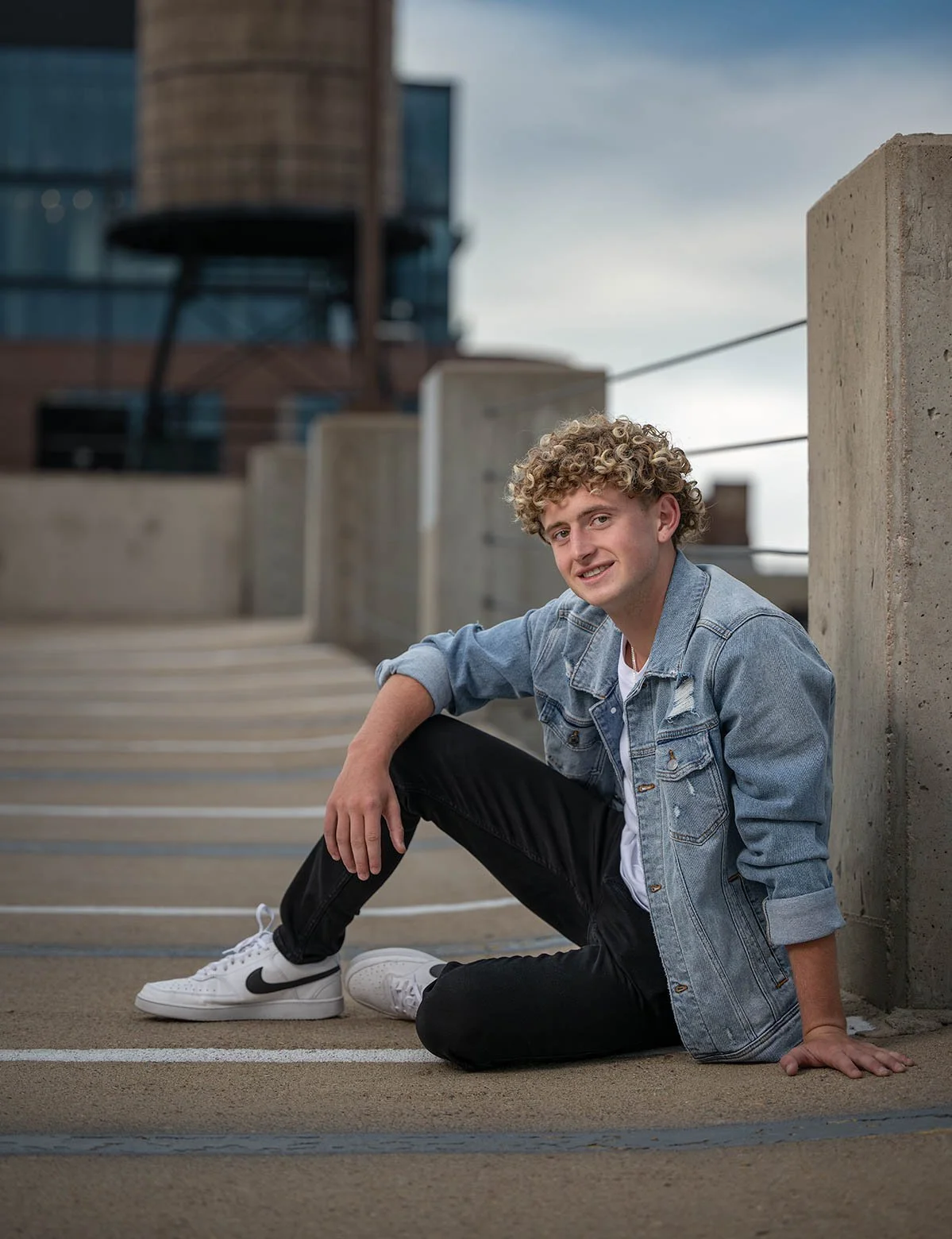 Young man with curly blonde hair wearing a denim jacket, white sneakers, and black pants, sitting on the ground in an urban setting with concrete structures and a building in the background.