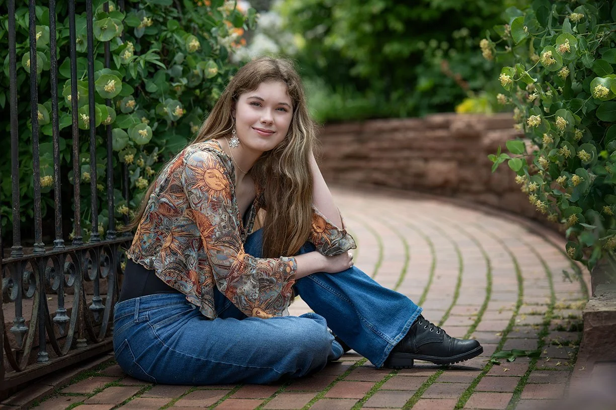 Natural senior portraits sitting on a brick pathway next to a black iron fence and green bushes with yellow flowers, smiling at the camera.