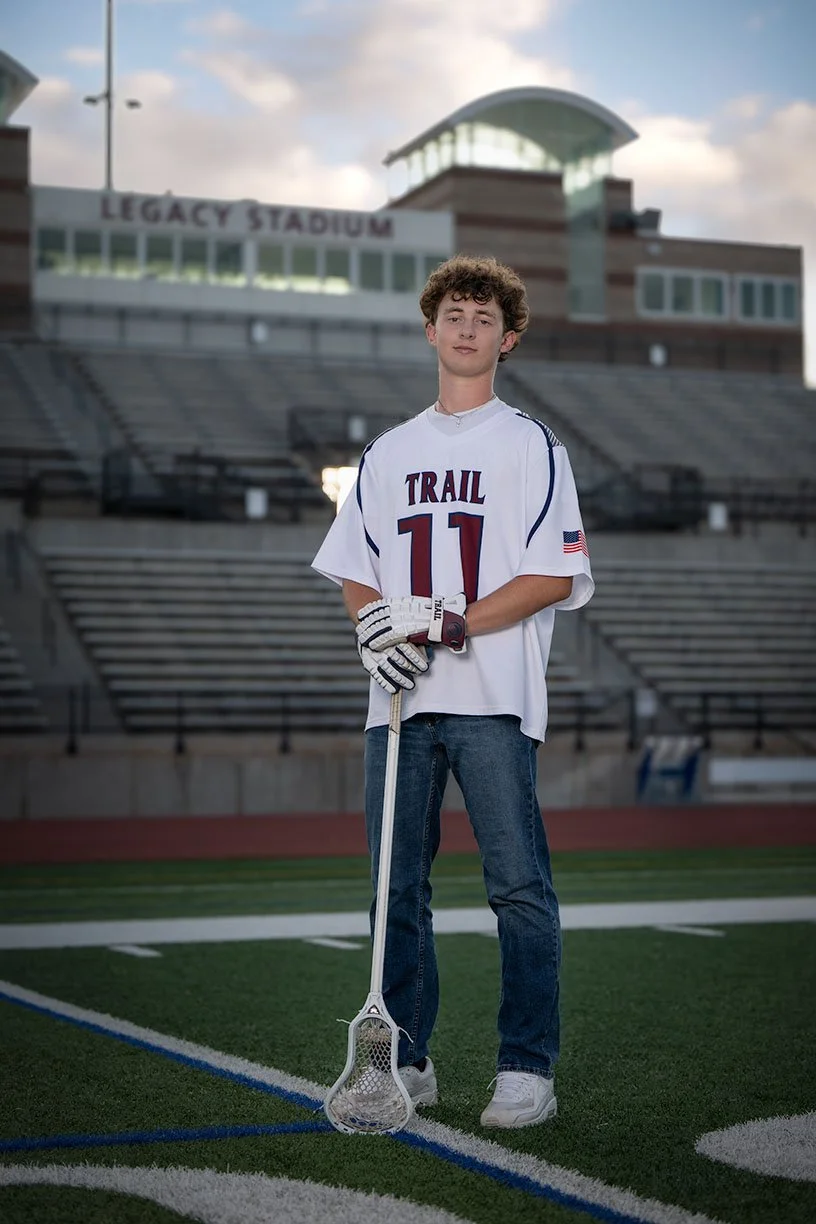 Sport senior portraits lacrosse jersey and gloves holding a lacrosse stick stands on a football field. In the background, there are empty stadium seats and a sign that says "Legacy Stadium."