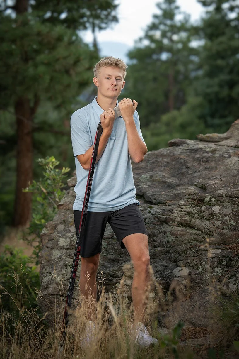 Senior portraits outdoors on rocks, holding a hockey stick over his shoulder, surrounded by trees and grass.