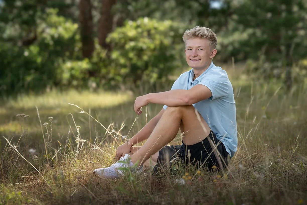 A young man sitting on grass in a park, smiling, wearing a light blue shirt, black shorts, and white sneakers, with trees in the background.