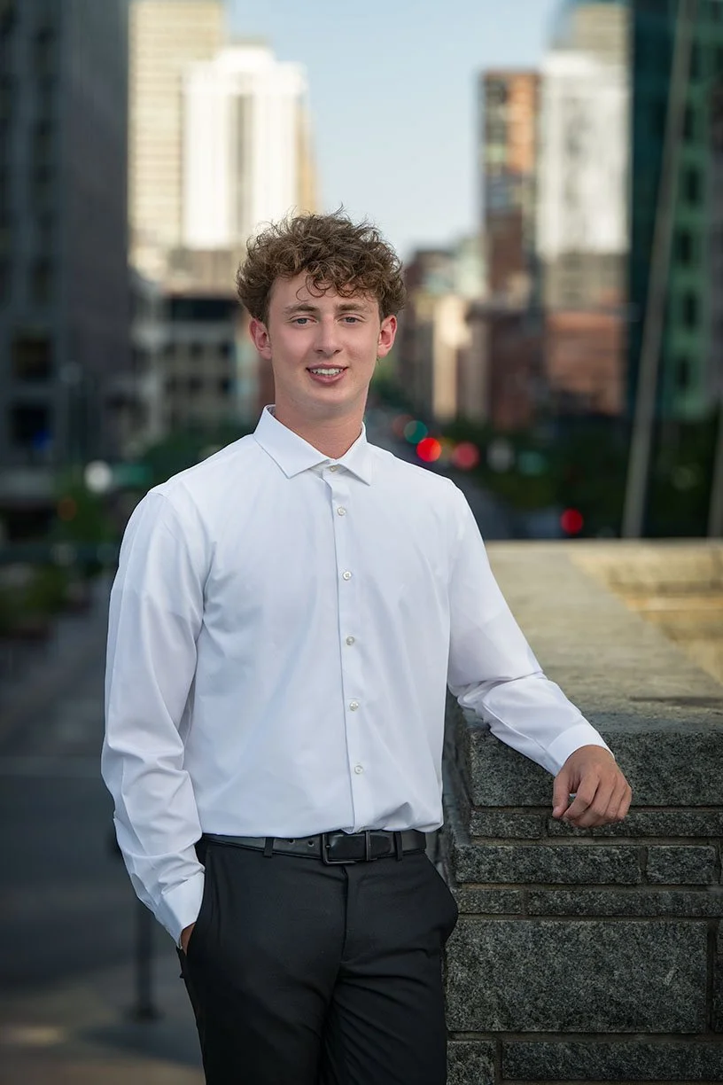 A young man with curly hair wearing a white dress shirt and black pants, standing outdoors on a city sidewalk, with a cityscape in the background.