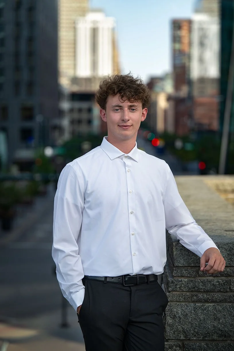 A young man with curly hair wearing a white dress shirt and black pants, standing outdoors in an urban setting with tall buildings in the background.