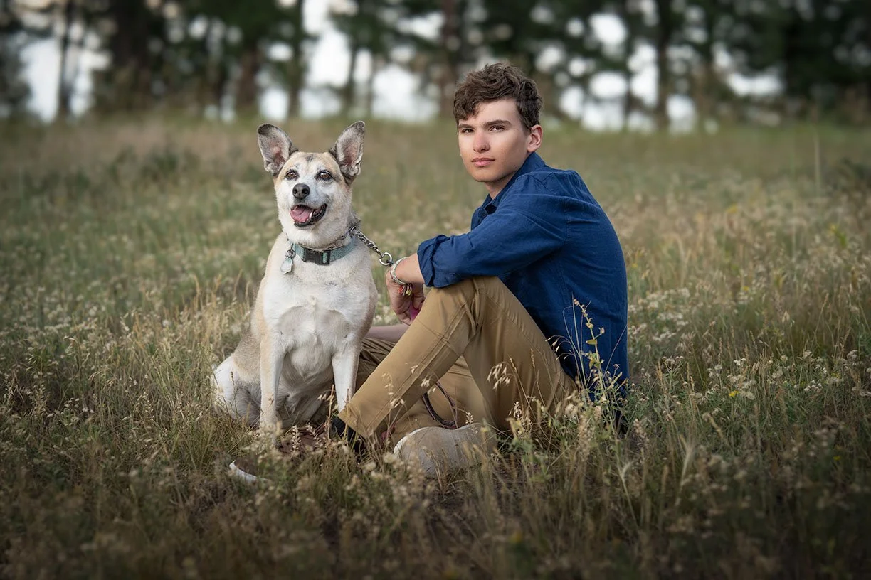 A young man in a blue shirt and tan pants sitting on grass with a mixed breed dog in a field of grass and wildflowers, with trees in the background.
