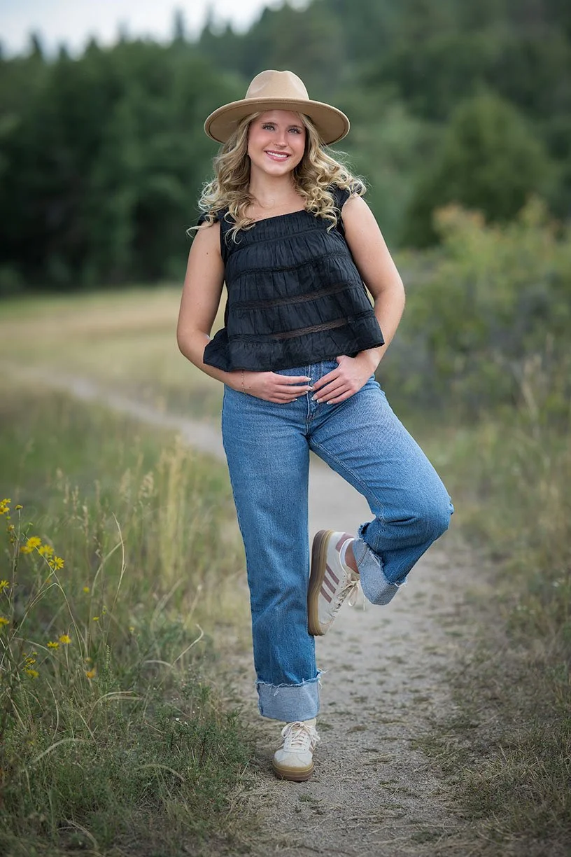 A woman wearing a wide-brimmed hat, black ruffled top, and blue jeans standing on a dirt path outdoors in a natural setting.