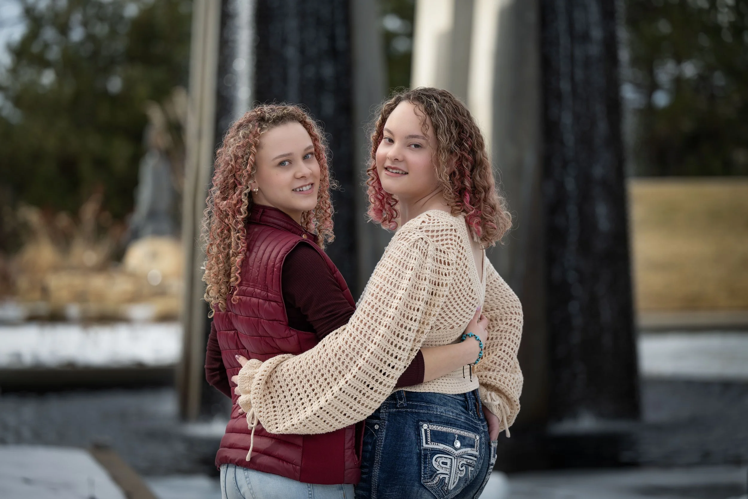 Two young women with curly hair standing outdoors near a fountain, embracing and smiling at the camera.