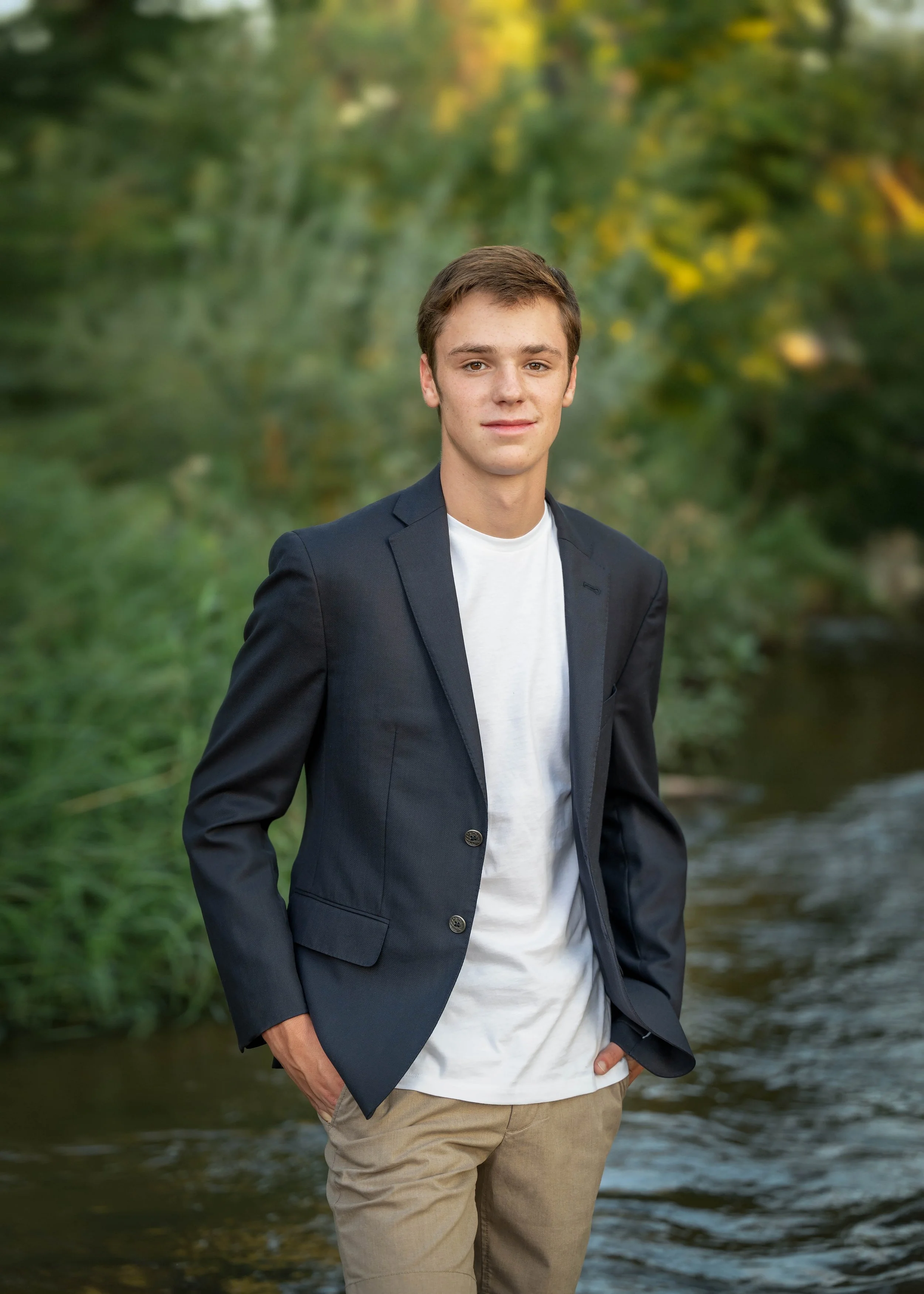A young man in a navy blazer, white t-shirt, and beige pants standing outdoors by a stream with green trees in the background.