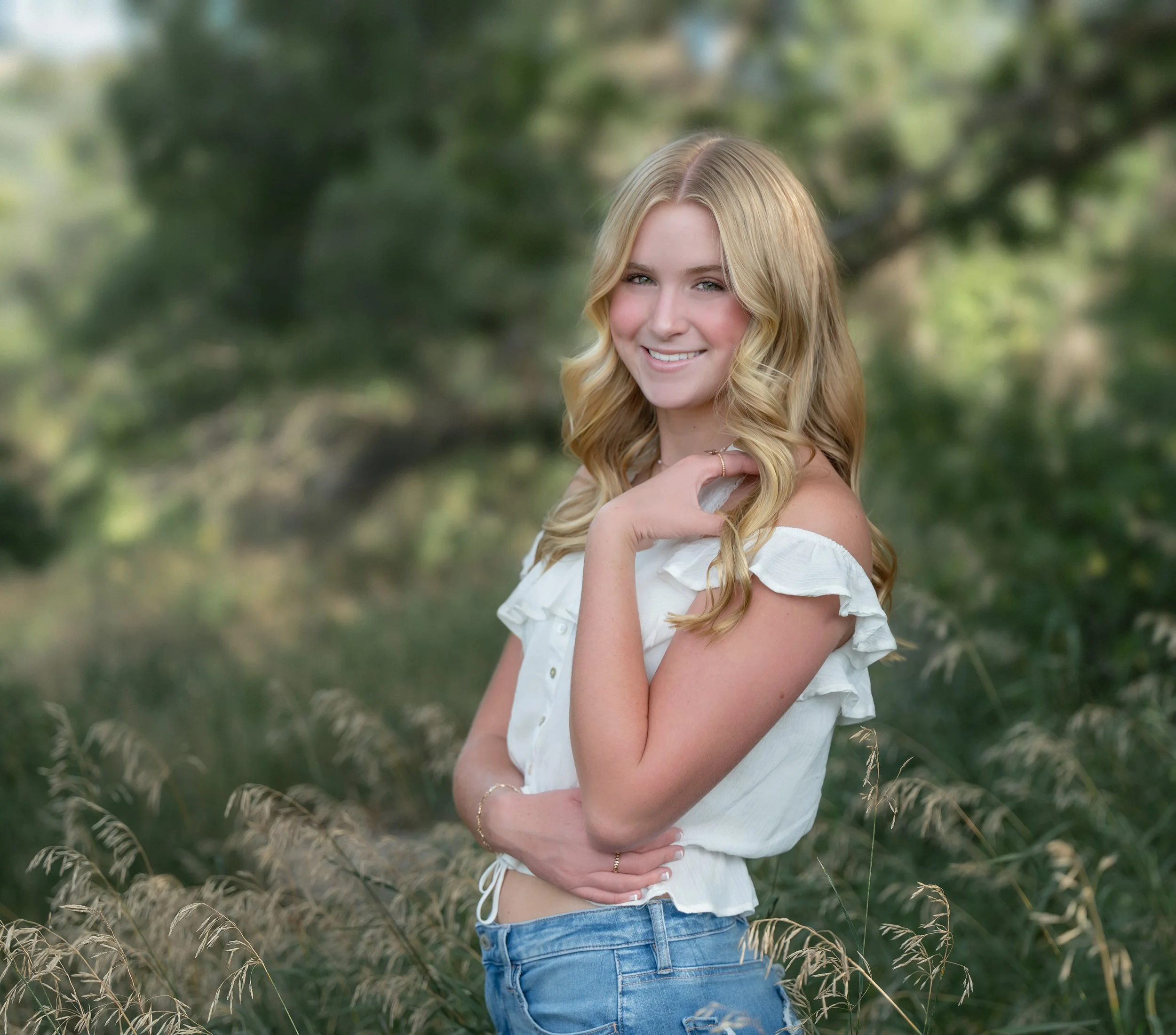 Young woman with blonde hair wearing a white off-shoulder top and blue jeans standing outdoors in a natural setting with trees and tall grass.