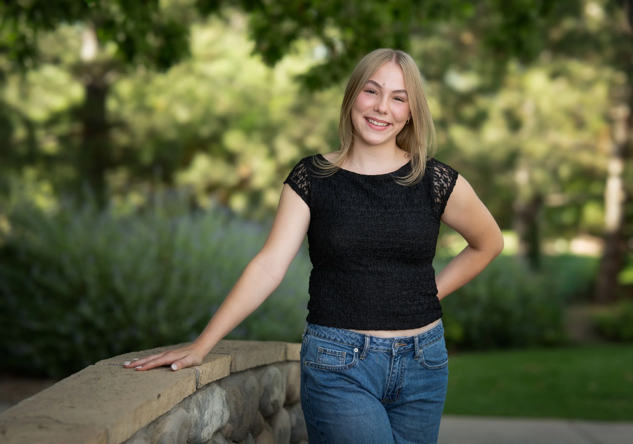 A young woman with blonde hair, wearing a black lace top and blue jeans, smiling and standing outdoors by a stone wall in a park or garden setting with trees and greenery in the background.