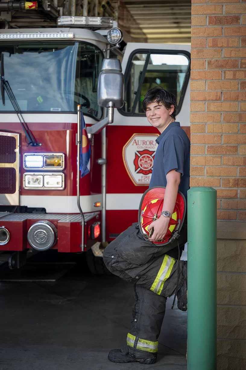 Young firefighter in uniform leaning against a green post, smiling, holding a red firefighter helmet, with a fire truck in the background.