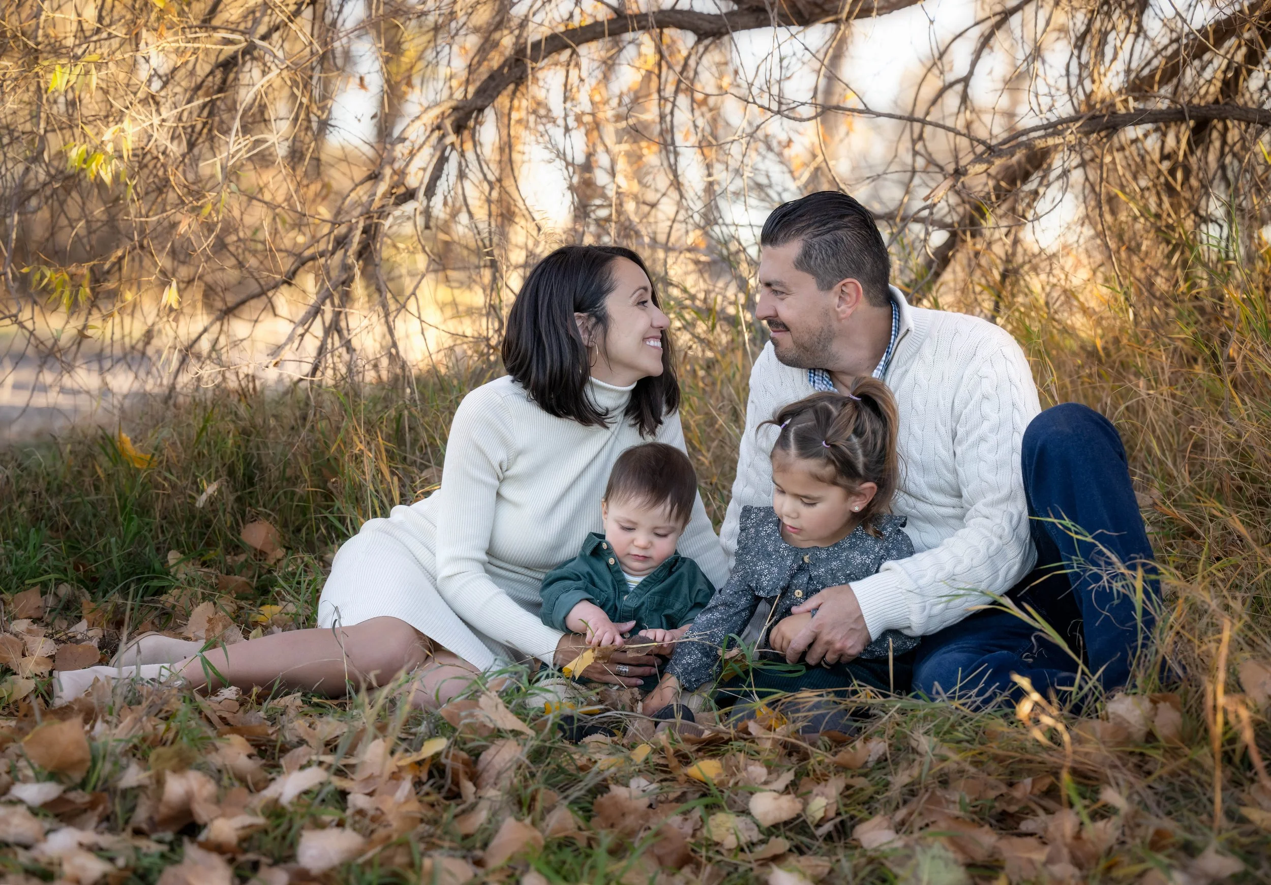 A family of four, two adults and two young children, sitting on the ground surrounded by fallen autumn leaves in a wooded area with autumn-colored trees. They are smiling and looking at each other.