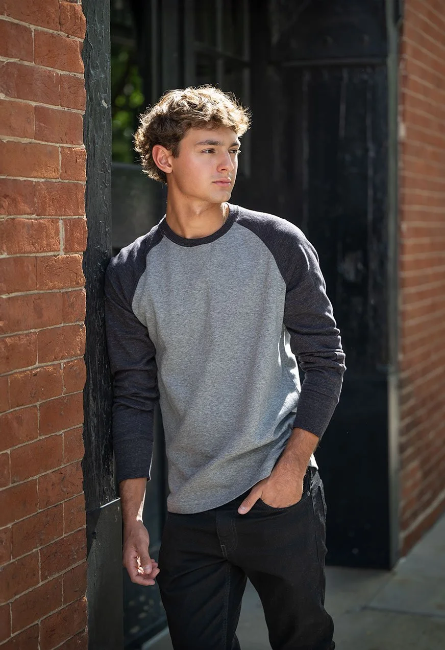 A young man with curly brown hair wearing a gray and black long-sleeve shirt and black jeans, leaning against a brick wall outside, looking to the side.